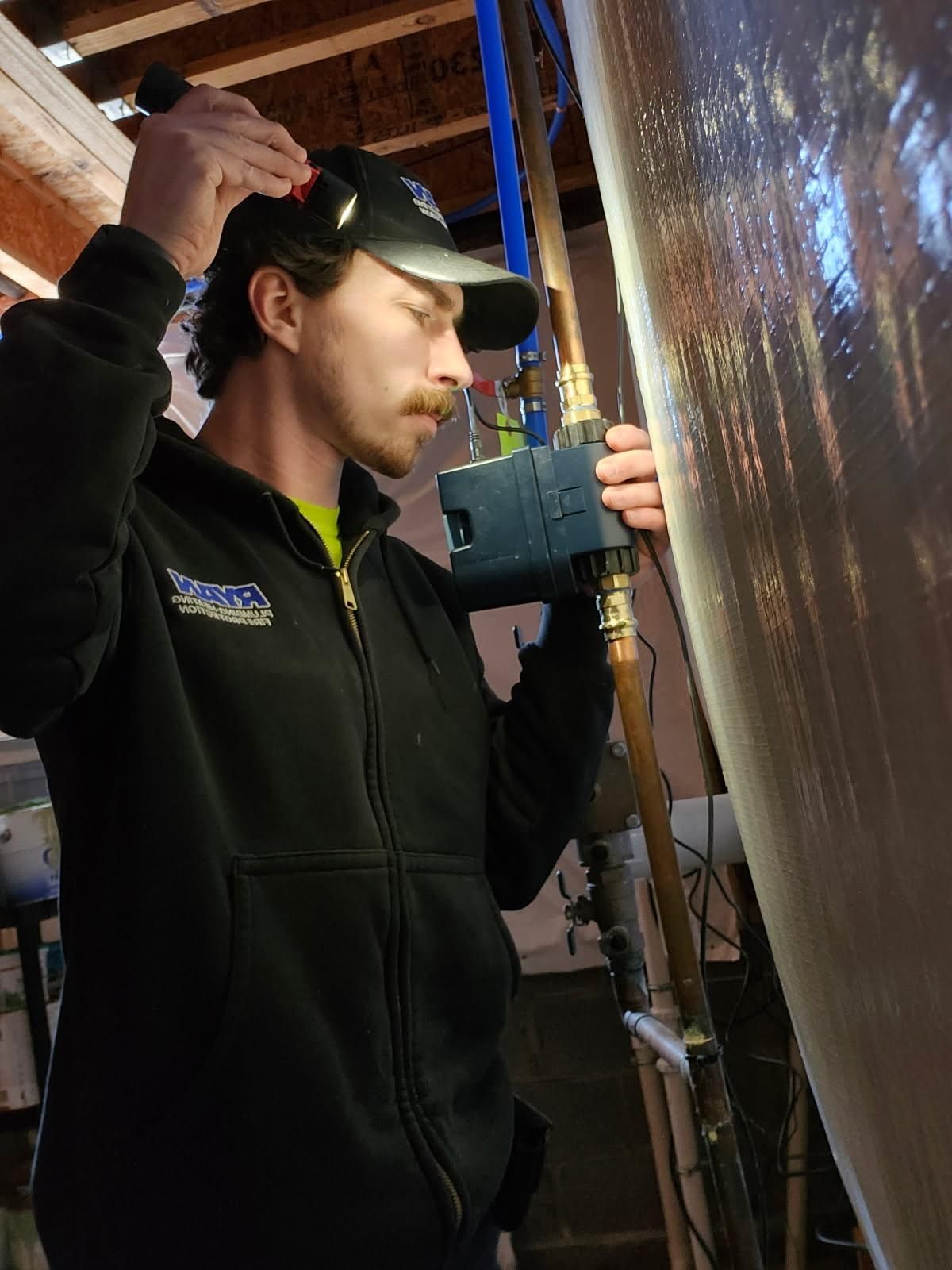 Man in a black jacket, hat, and flashlight inspecting water system near a large tank.