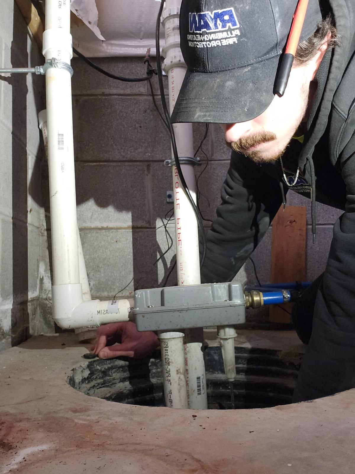Man inspecting a sump pump in a basement, wearing a hat, looking down at it.