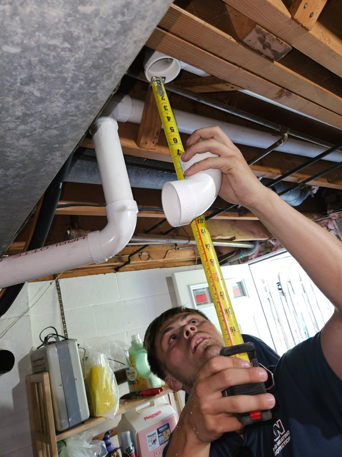 A man in a basement measures PVC pipe with a tape measure, preparing for plumbing work.