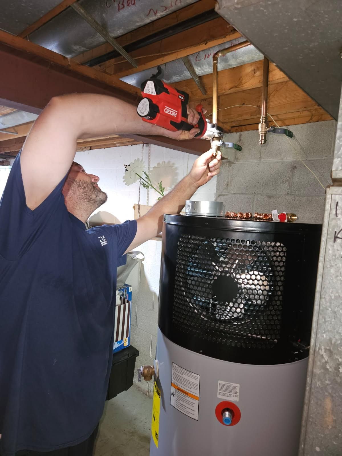 A man in a blue shirt works on copper pipes near a water heater in a basement setting.