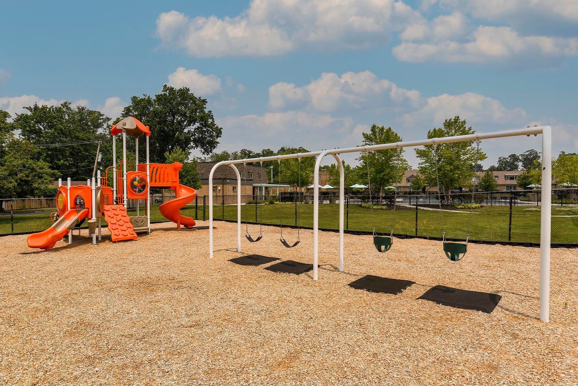 Orange playground equipment with swings on wood-chip ground, fence and apartment buildings in the background.