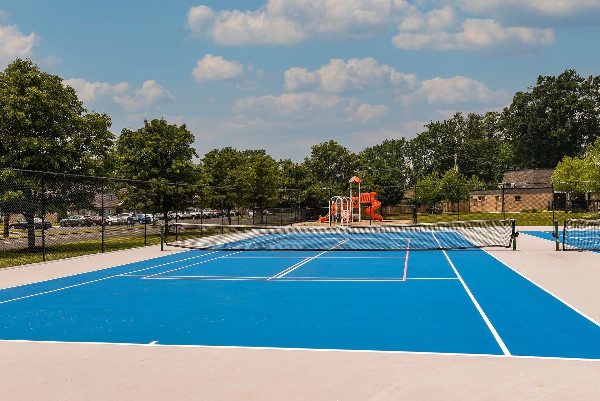 Blue outdoor sport court with a net, fence, and surrounding trees.