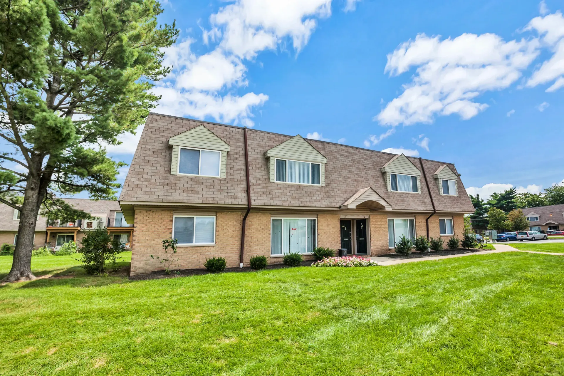 Exterior view of a brick apartment building with a green lawn, trees, and clear blue sky.