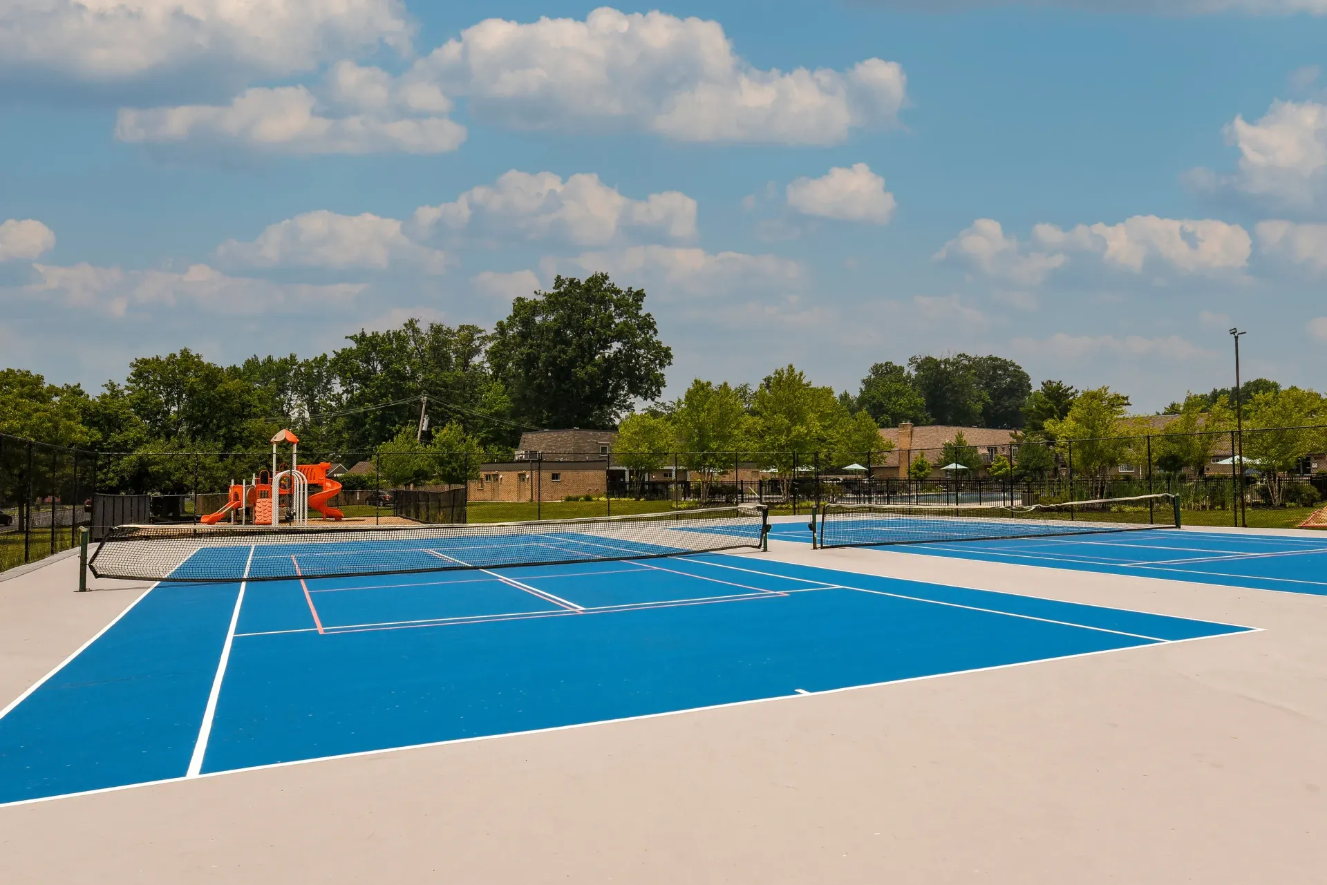 Outdoor blue tennis court with nets, fenced, and a playground in the background.