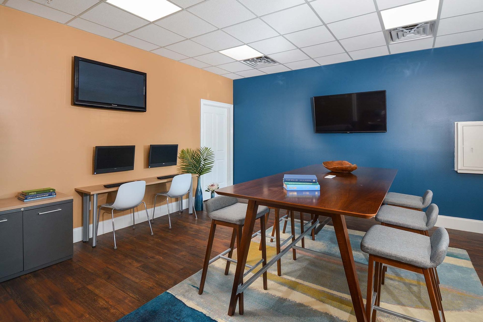 Community lounge with a long wooden table, gray chairs, computer workstations, and wall-mounted TVs.