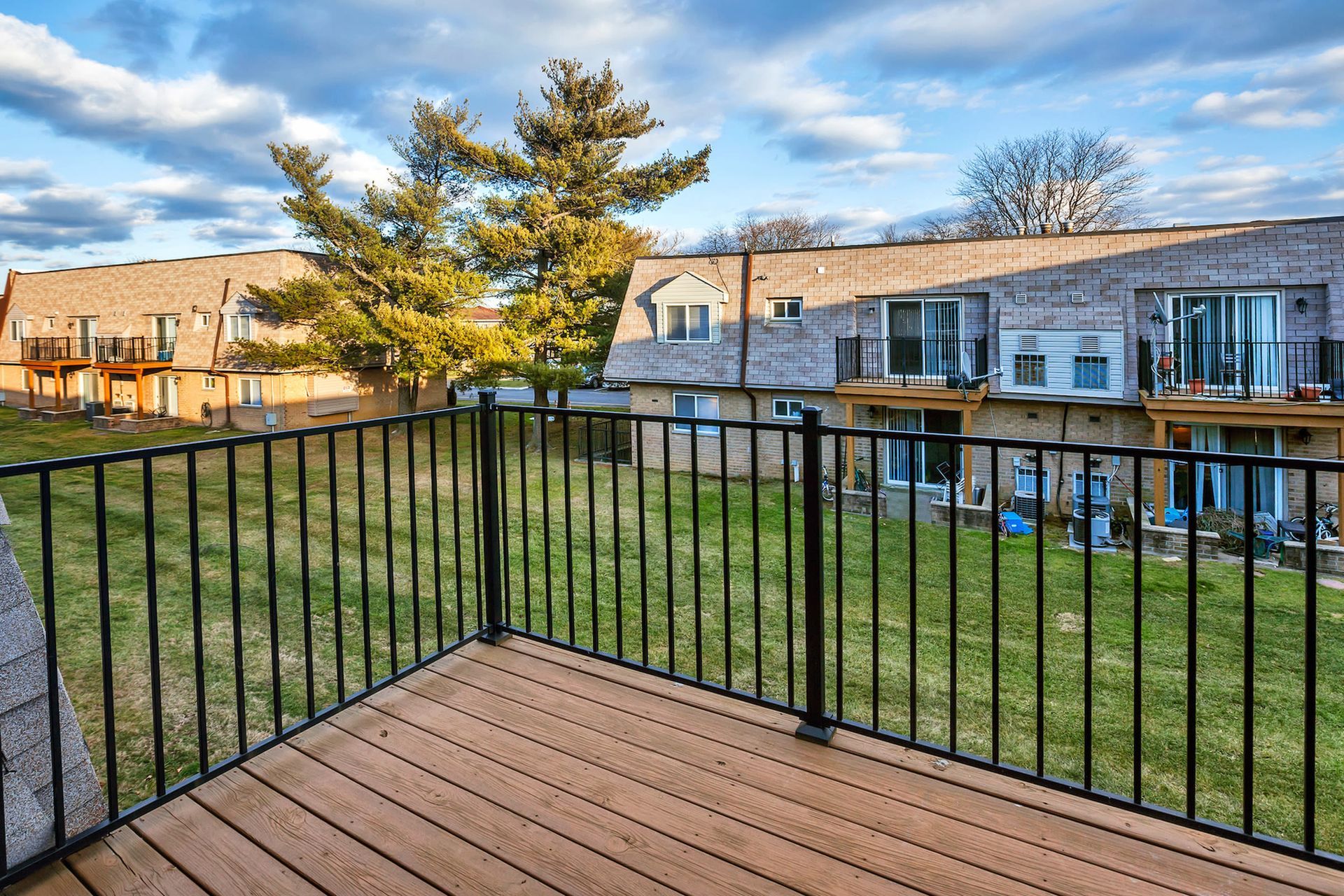 Balcony with a black railing and wooden deck overlooks a grassy courtyard and row of brick apartment buildings.