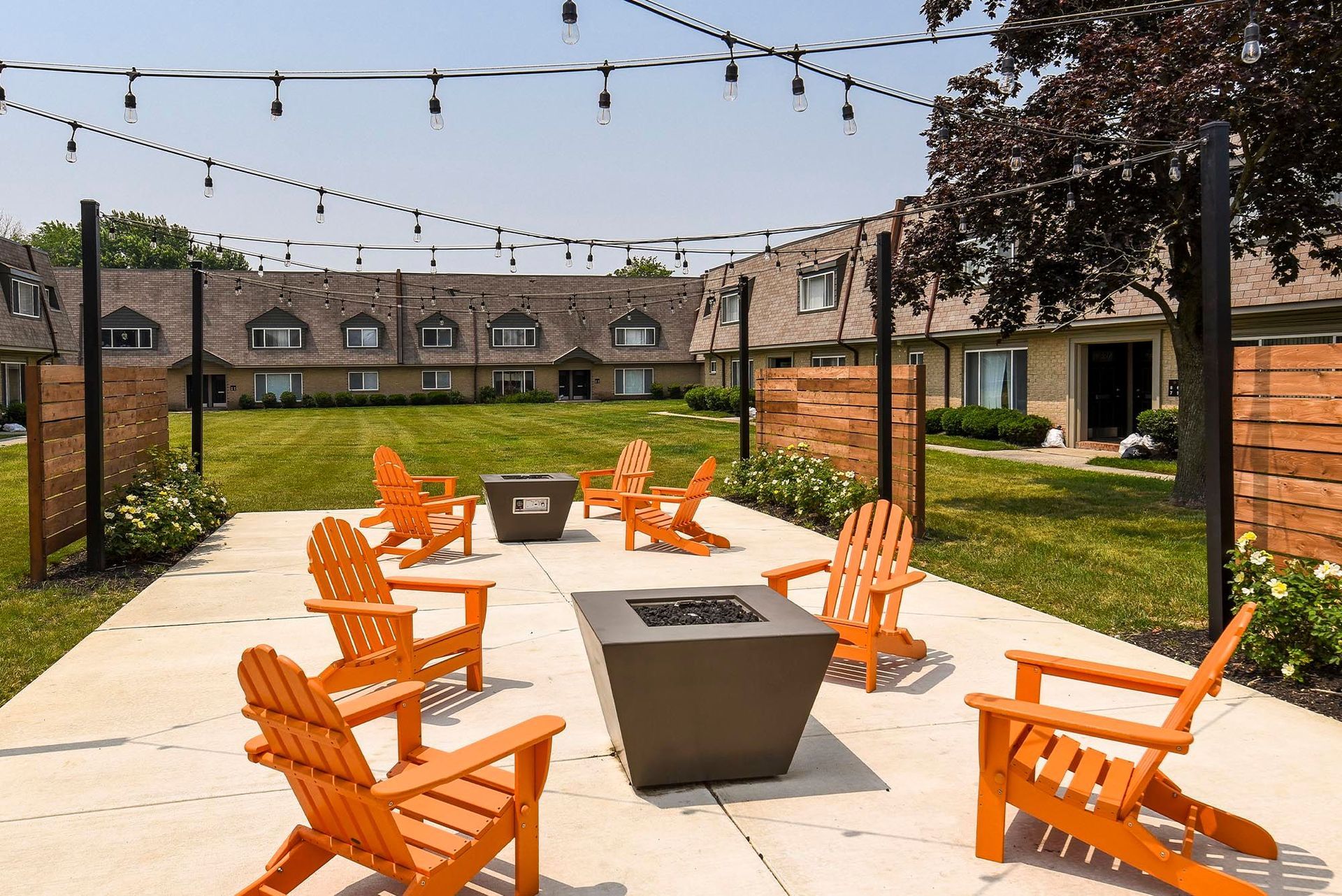 Outdoor communal seating area with orange Adirondack chairs around fire pits and string lights.