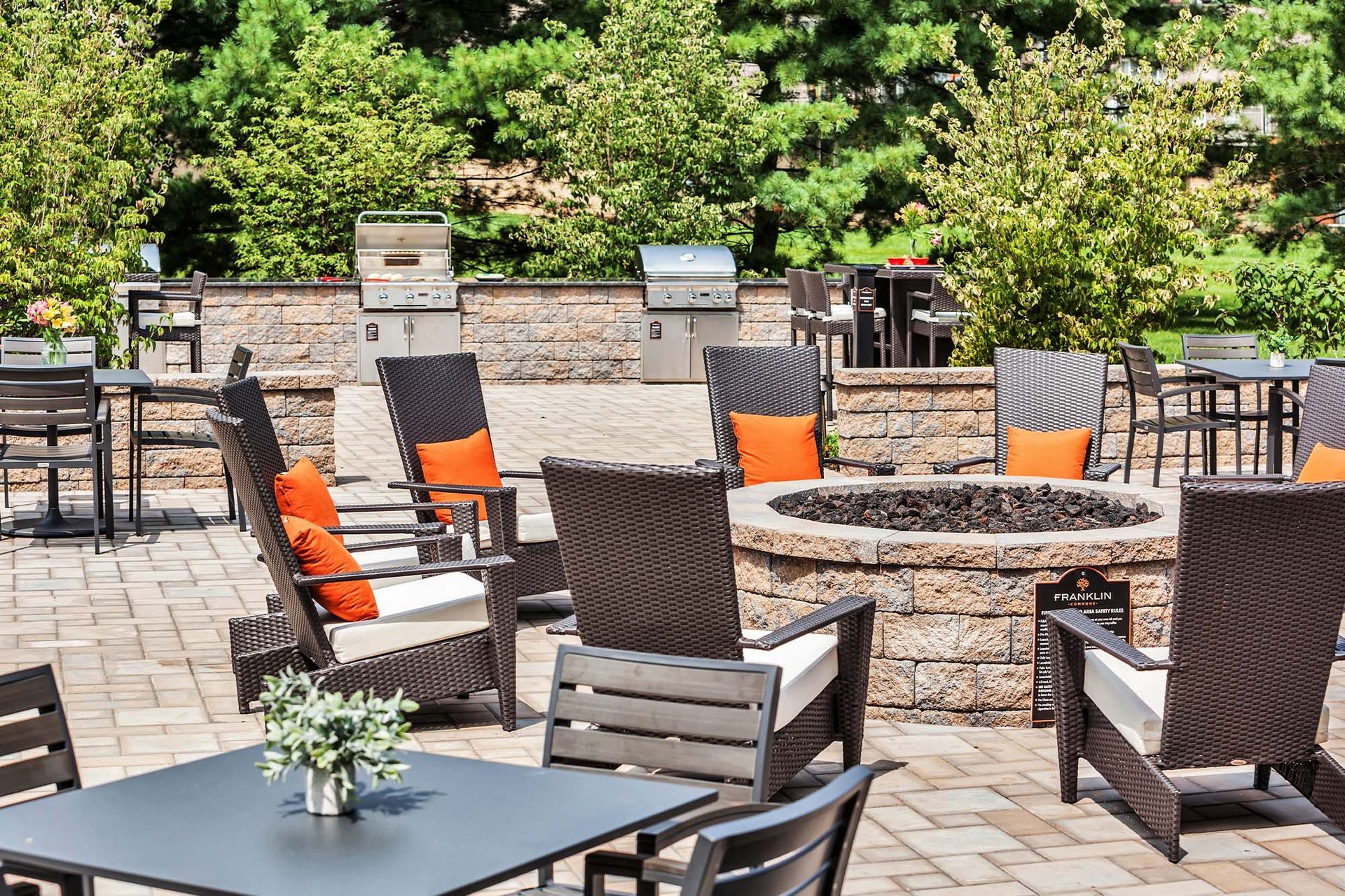 Outdoor community patio with a stone fire pit, wicker chairs with orange cushions, and grills.