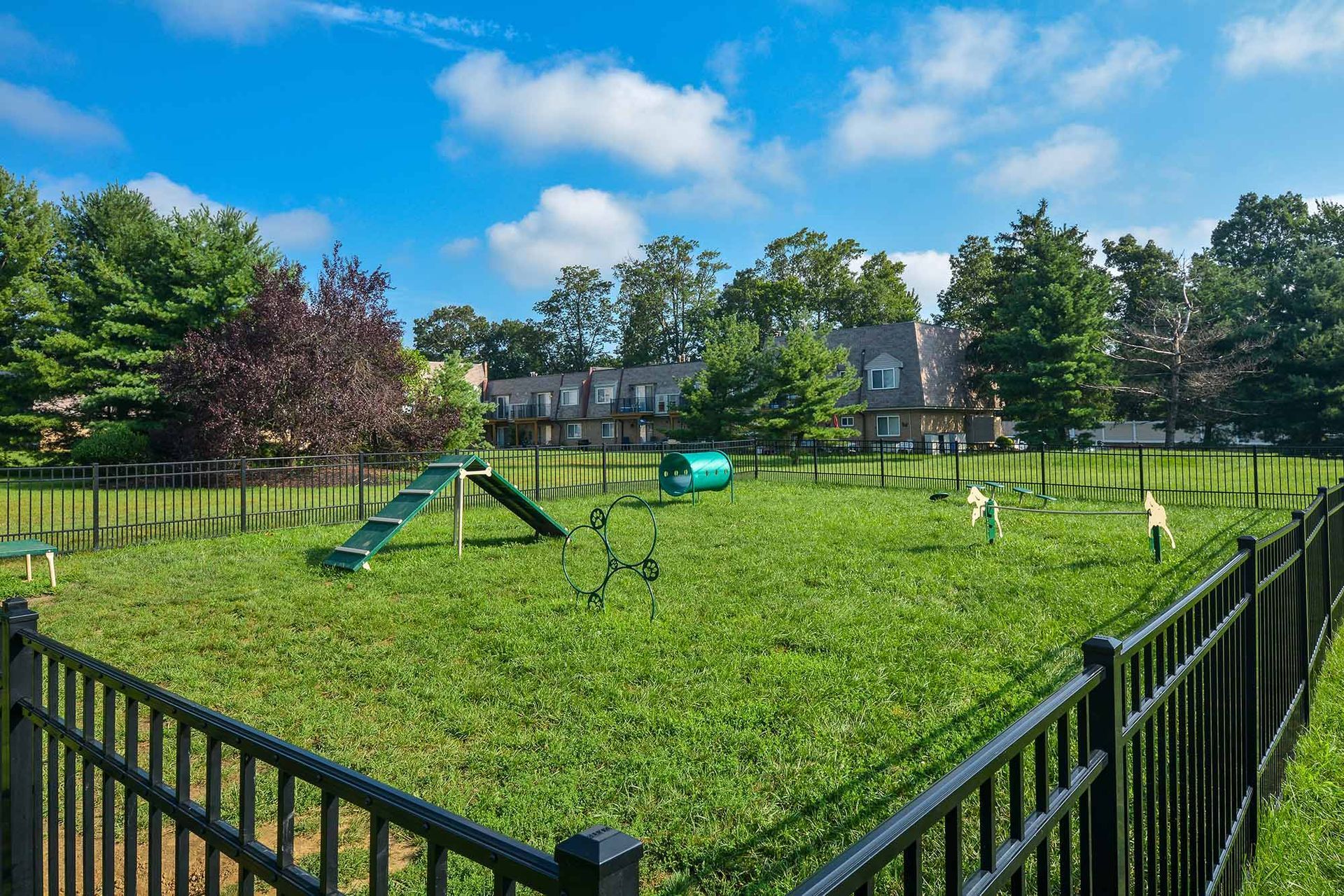 Fenced community playground with grass and play structures, with apartment buildings in the background.