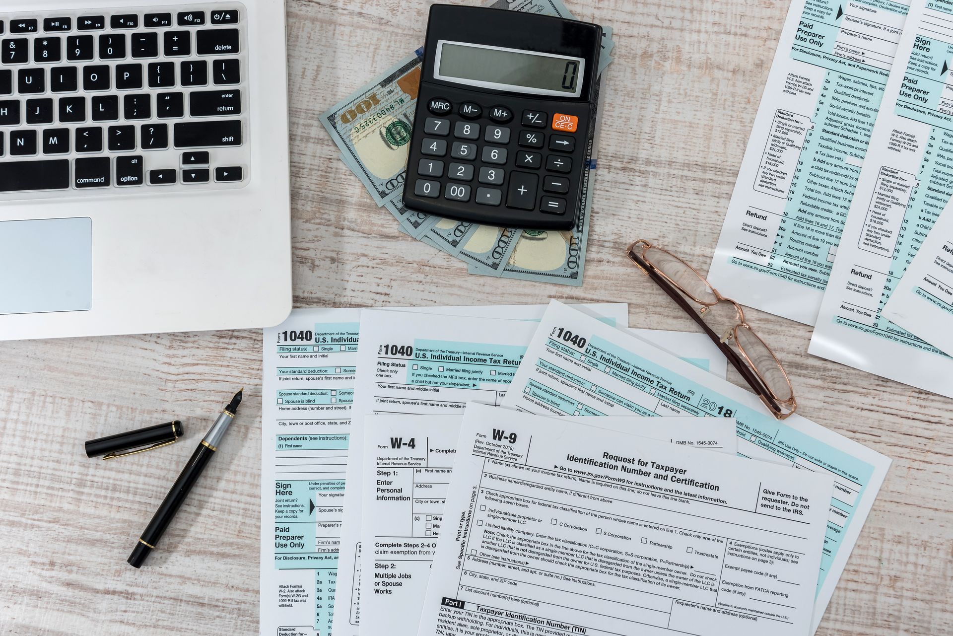 Calculator, cash, and US tax forms on a desk beside a laptop, pen, and glasses.