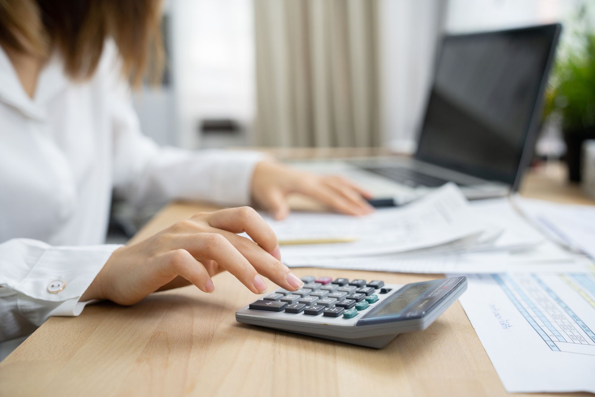Close-up of a woman's hands using a calculator and laptop for tax preparation at a home office.