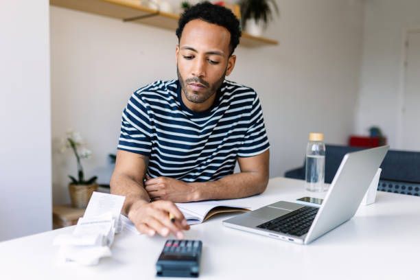 A young adult man with a laptop checking taxes sitting at his living room table.