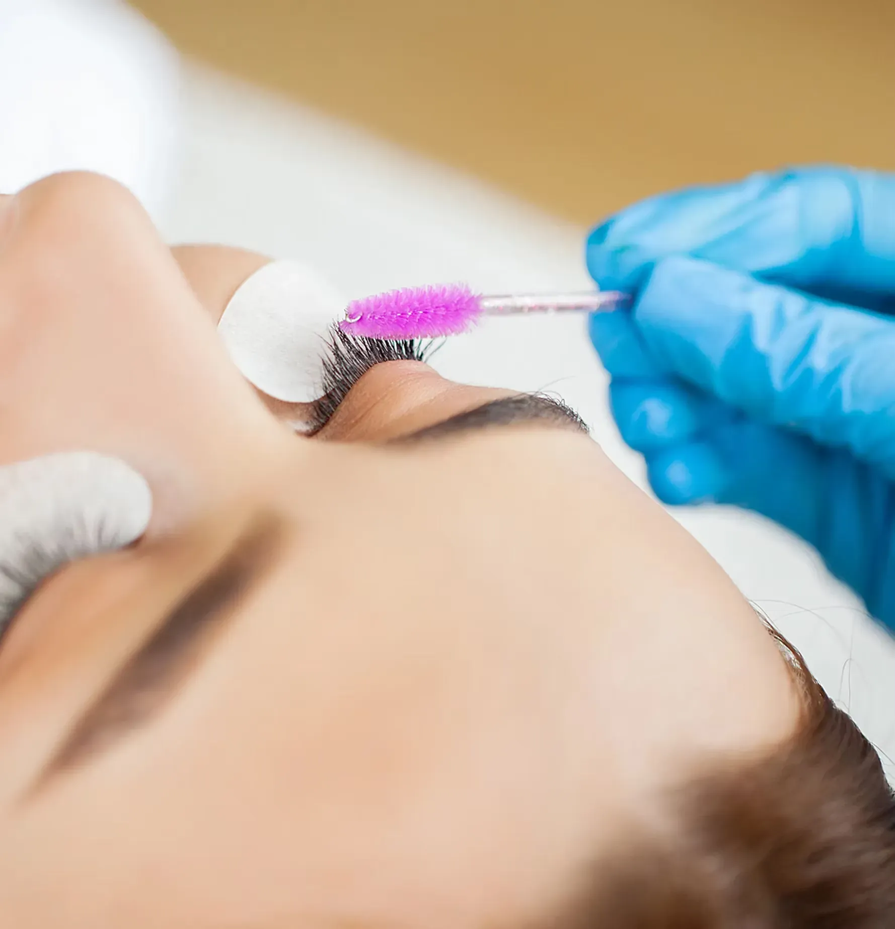 A close-up view of a professional using a pink mascara wand to groom a client's eyelashes during a lash treatment.