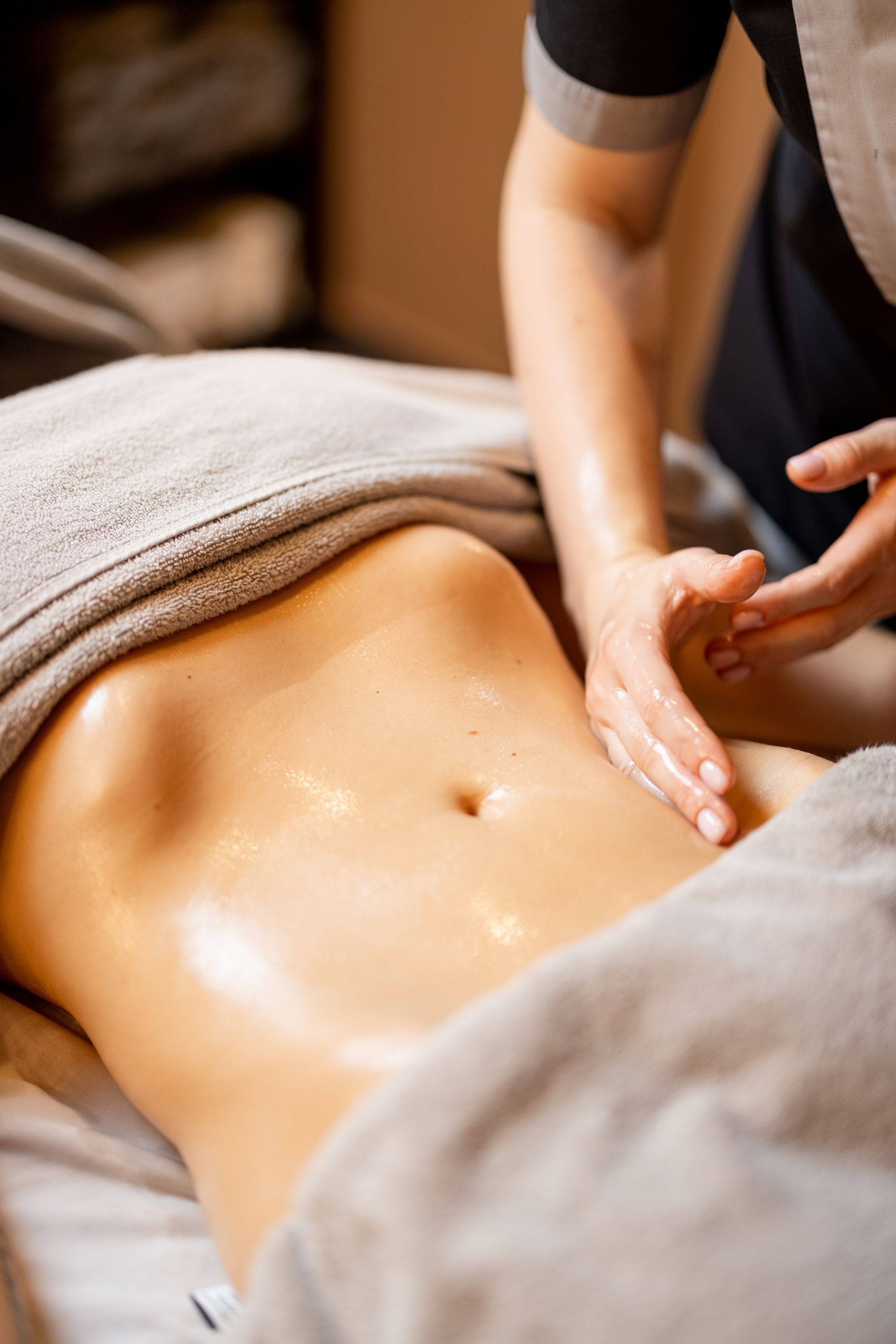 A massage therapist performs an abdominal massage on a person lying under a grey towel in a spa setting.