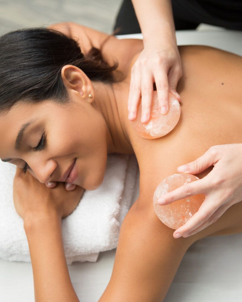 A person receives a back massage with warm, circular pink salt stones while relaxing on a white towel.