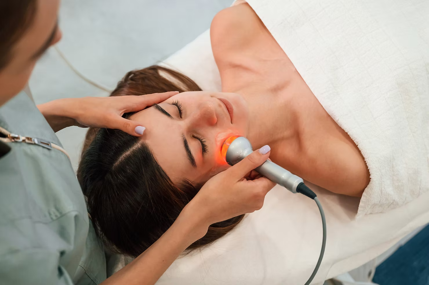 A professional aesthetician uses a handheld red light therapy device on a client’s face during a spa treatment.