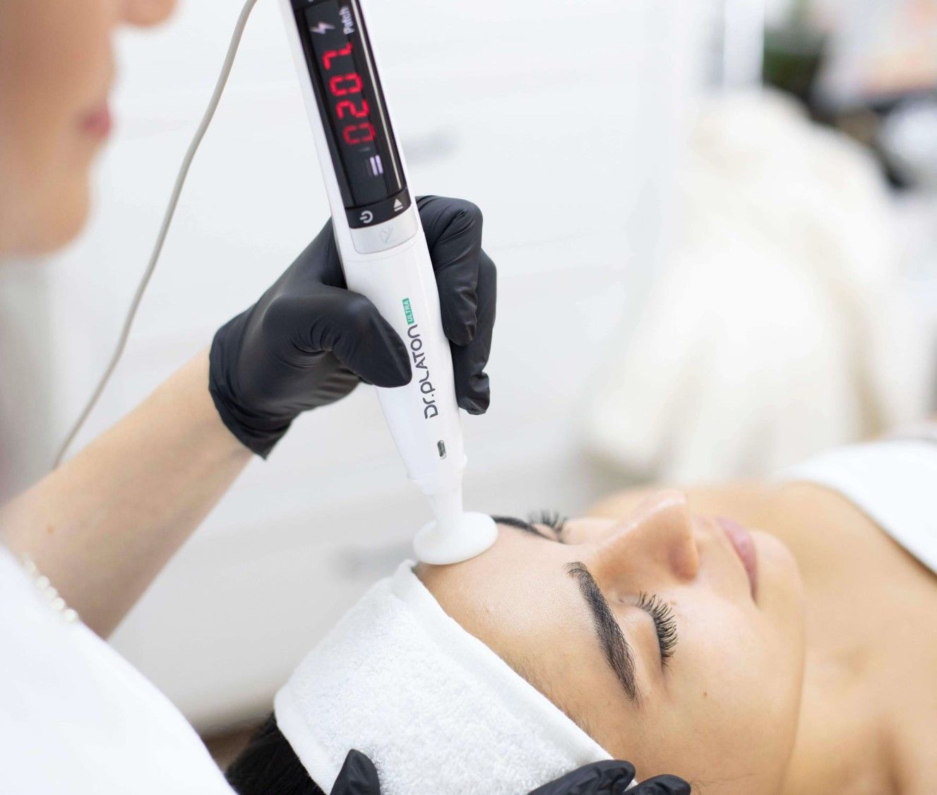 A professional practitioner in black gloves uses a cosmetic device on a person’s forehead in a spa setting.