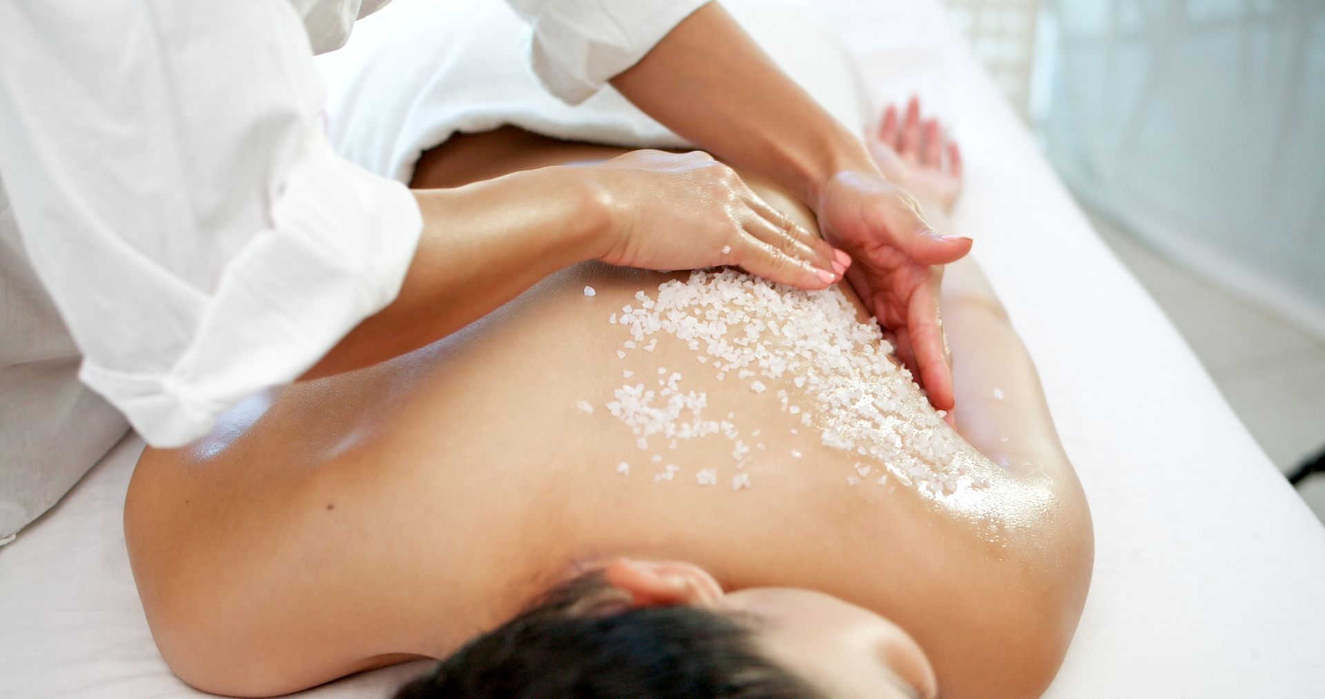 A massage therapist performs a salt scrub exfoliation on a client's back at a spa.