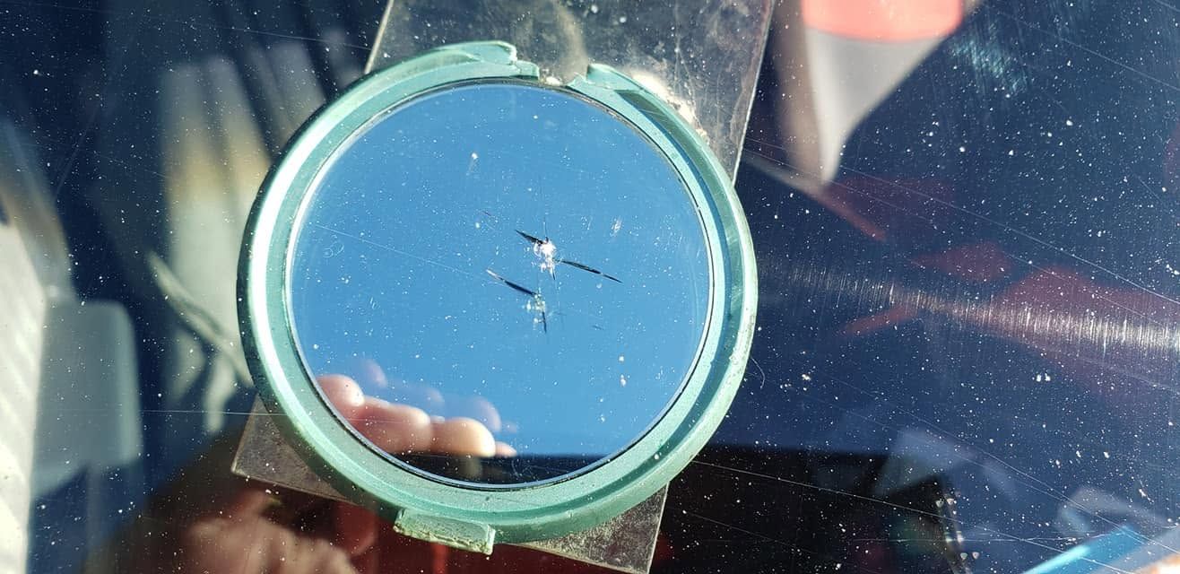 Round blue window or mirror reflecting a hand, seen through a car windshield with sun glare.