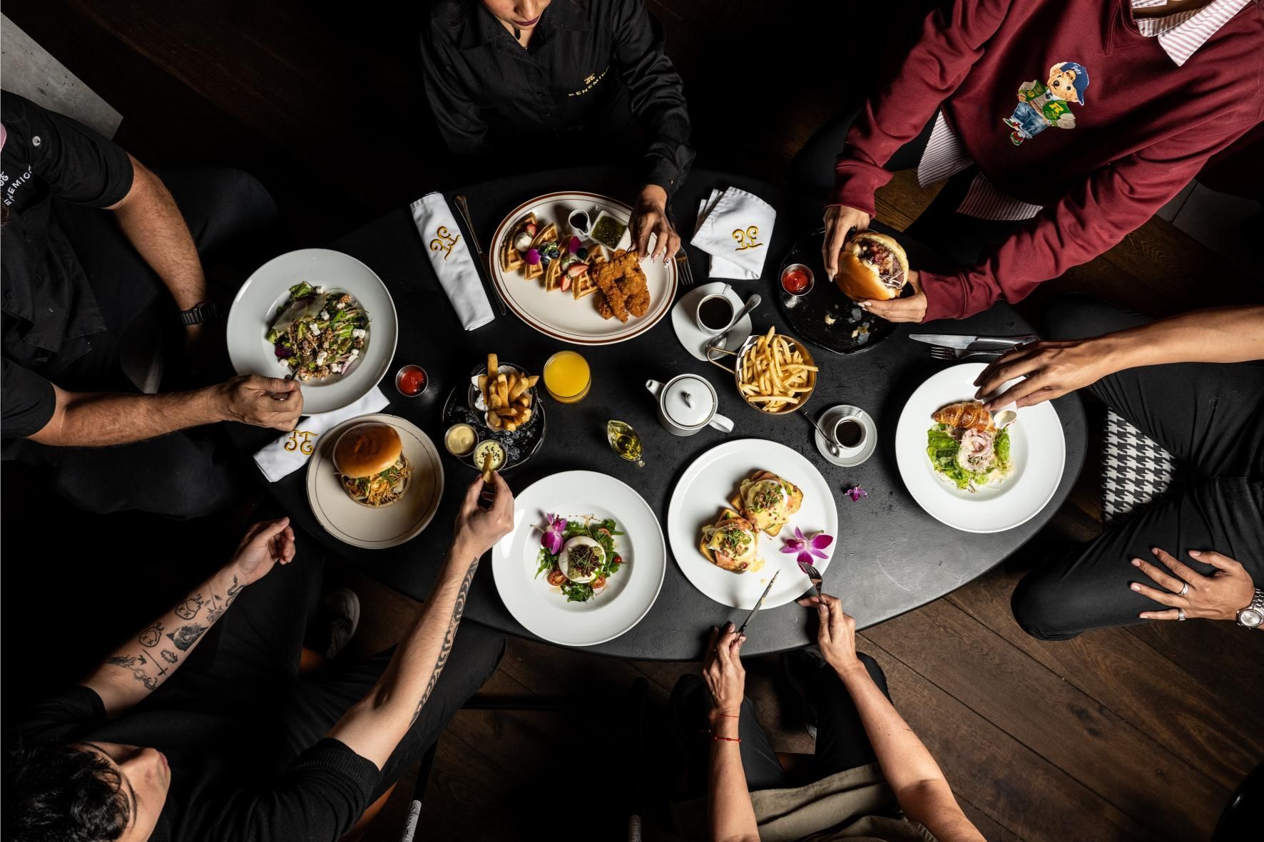 Personas alrededor de una mesa redonda comiendo un brunch: platos de comida, bebidas, manos extendidas. Ambiente oscuro.