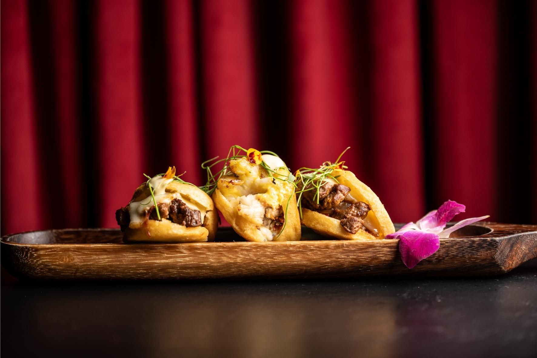 Tres mini tacos gourmet en una bandeja de madera, frente a una cortina roja.