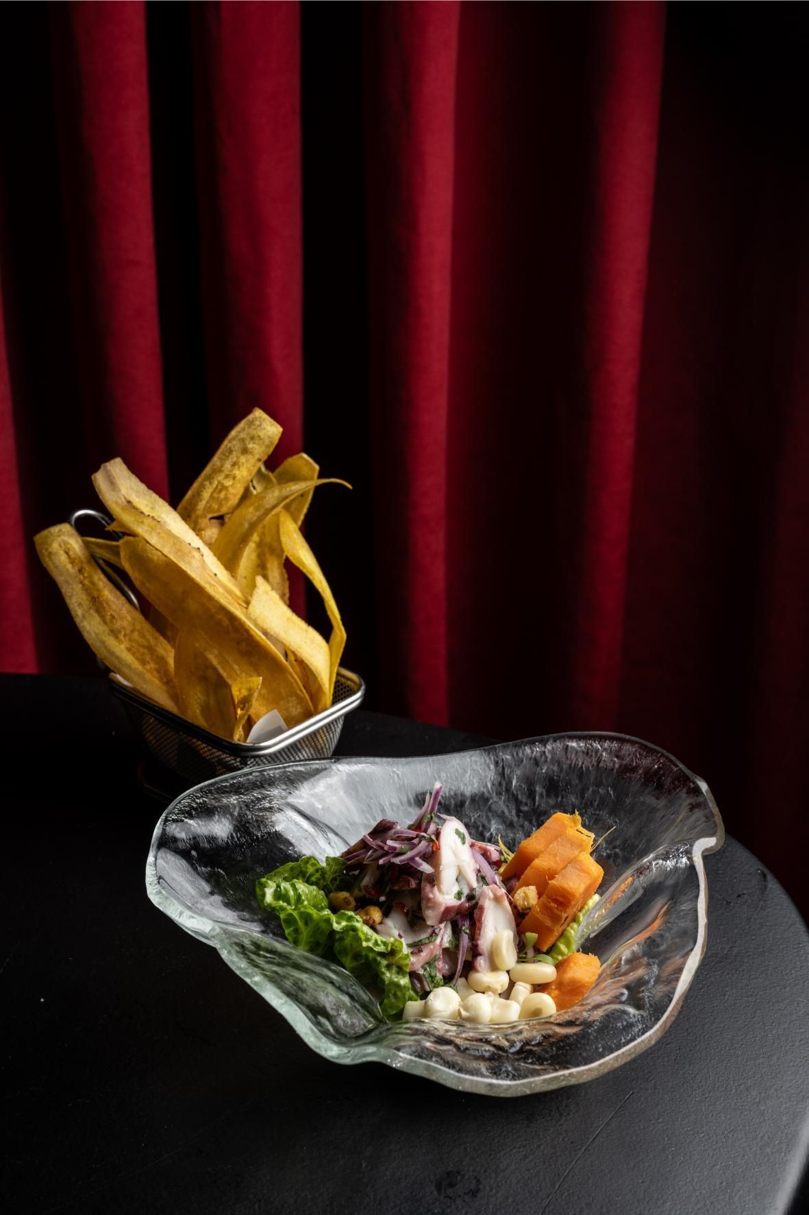 Ceviche con plátanos fritos en plato de cristal. Fondo de cortina roja.
