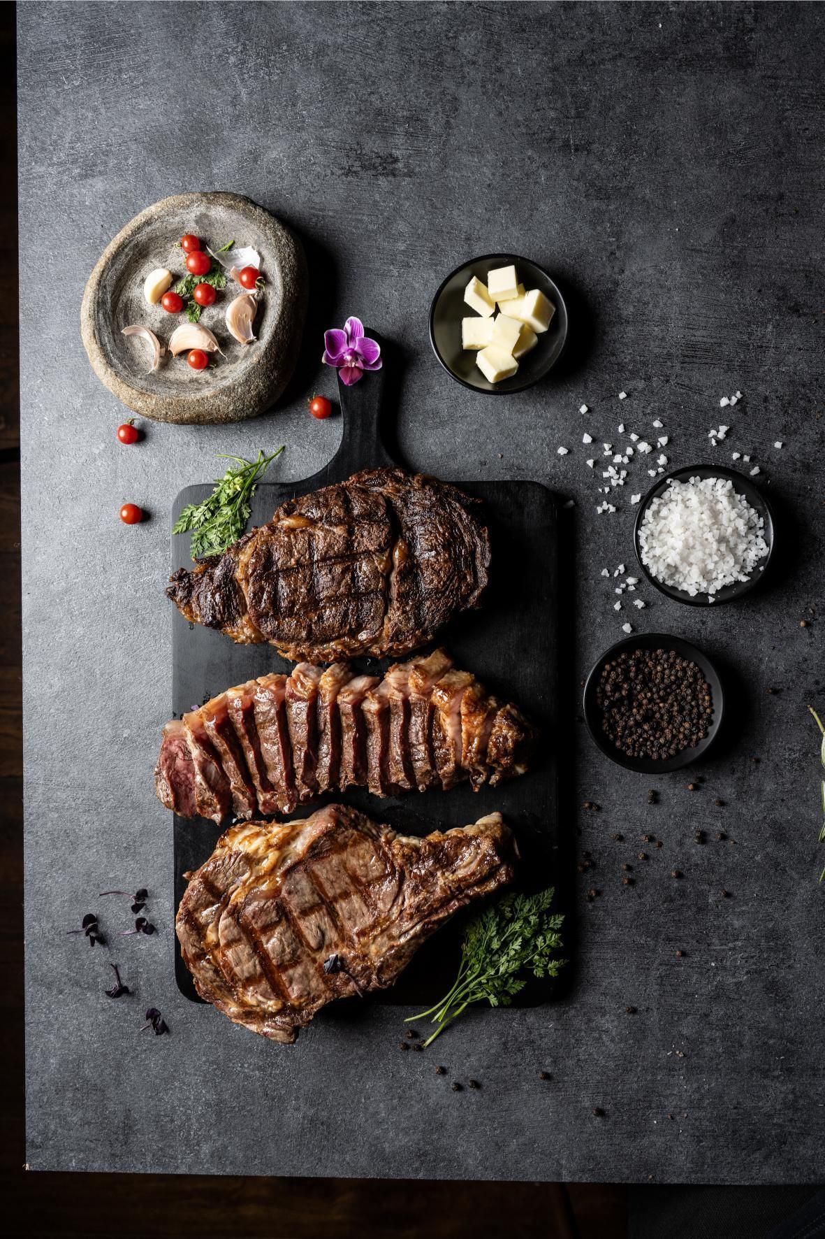 Tres filetes a la parrilla sobre una tabla de cortar negra con mantequilla, sal, pimienta, ajo y tomates.