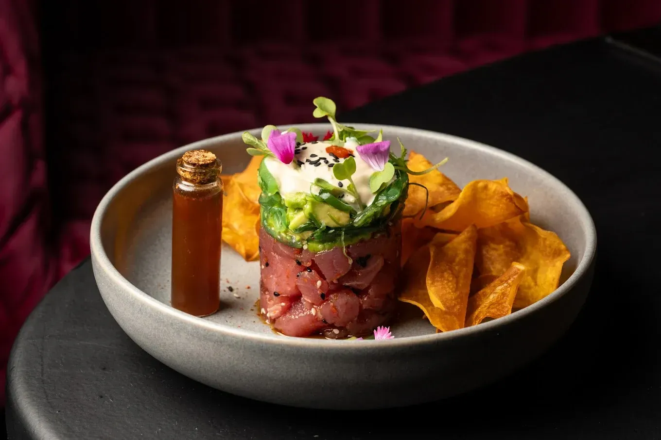 Torre de tartar de atún con aguacate, algas, patatas fritas y salsa sobre plato gris.