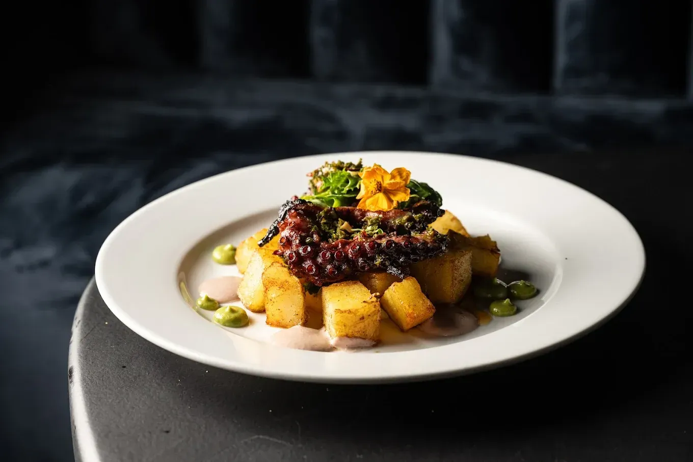 Papas doradas cortadas en cubos, cubiertas con salsa oscura, hierbas y flores comestibles en un plato blanco. Fondo oscuro.