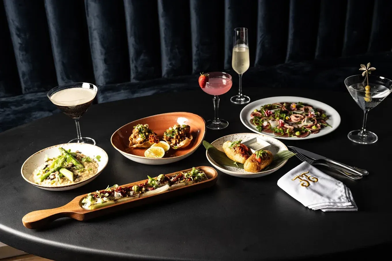 Una mesa con aperitivos y cócteles, incluido un martini, en un entorno poco iluminado.