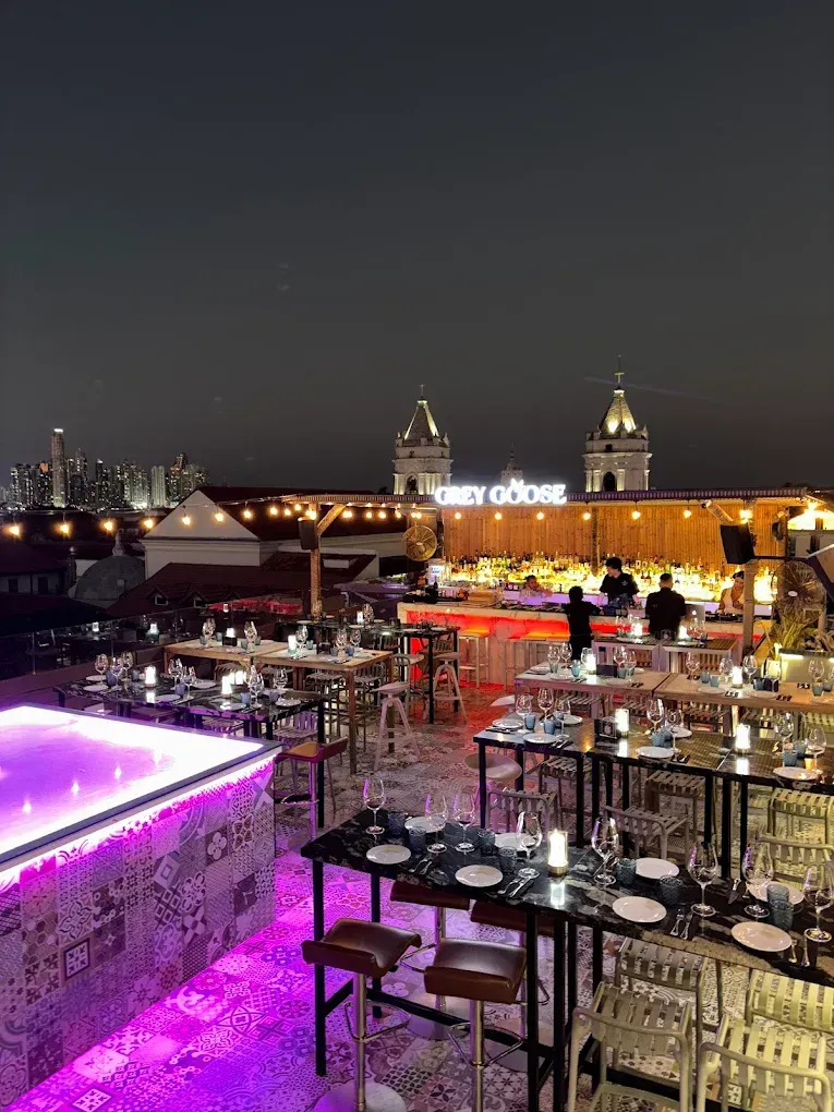 Bar en la azotea por la noche con mesas, luces y un cartel de Grey Goose al fondo.