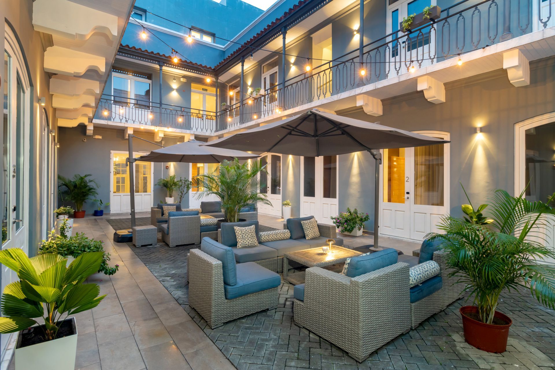 Patio exterior con muebles de ratán gris, sombrillas y plantas en macetas; iluminación nocturna.
