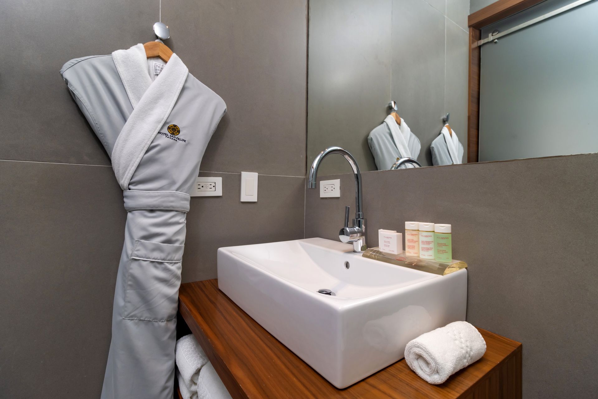 Baño con paredes grises, lavabo rectangular blanco sobre encimera de madera, albornoz colgado y espejo.