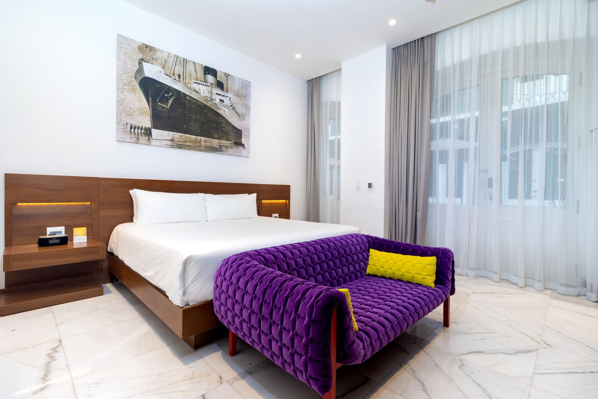 Dormitorio con cama, sofá morado y un barco en la pared. Suelo de mármol y cortinas transparentes.