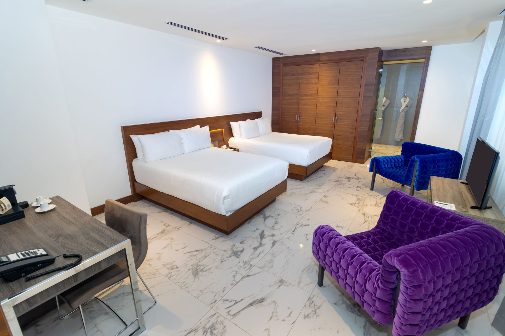 Habitación de hotel con dos camas, escritorio, sillones morados y azules y un armario.