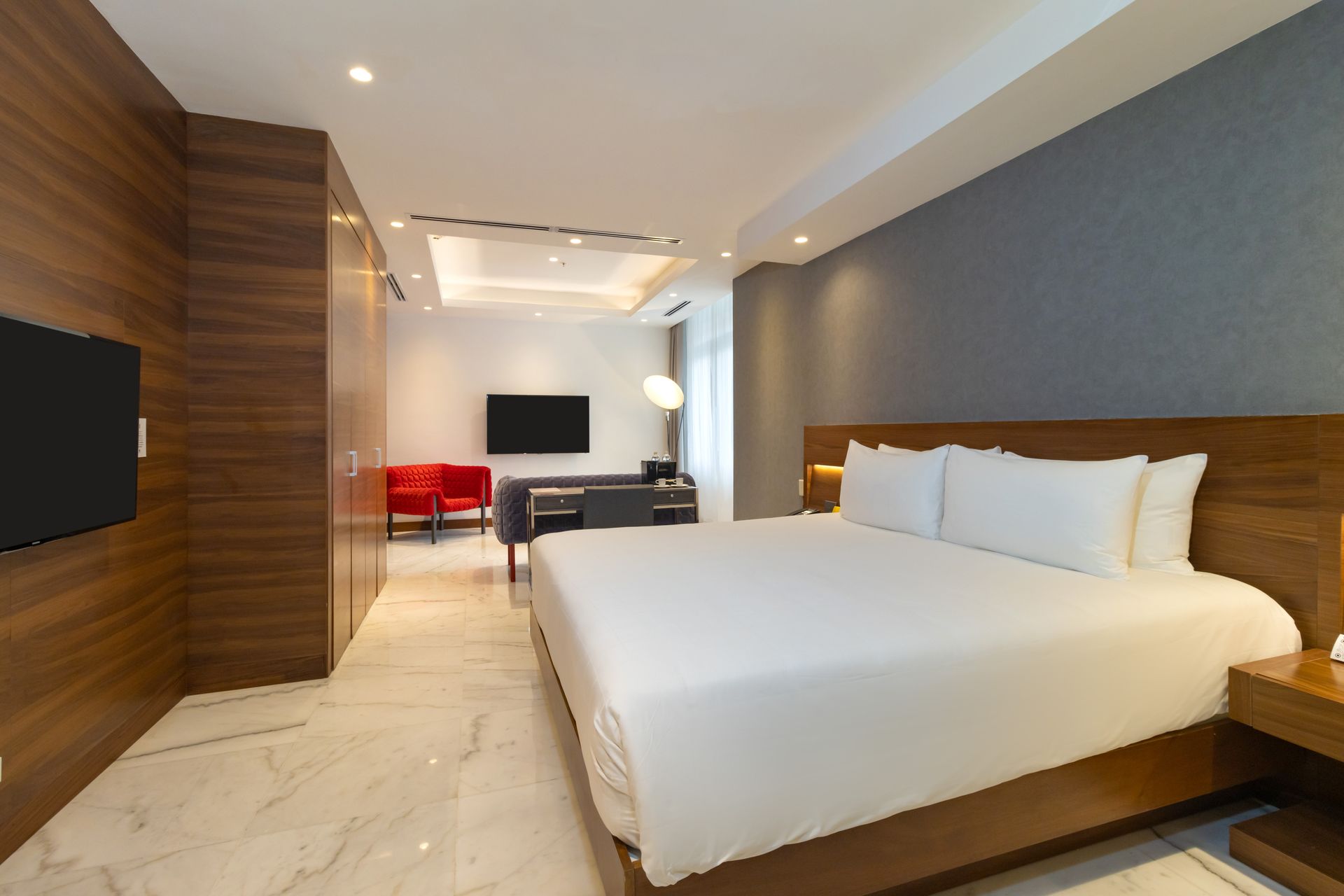 Habitación de hotel con cama grande, televisor, zona de estar y detalles en madera. Combinación de colores blanco y gris.