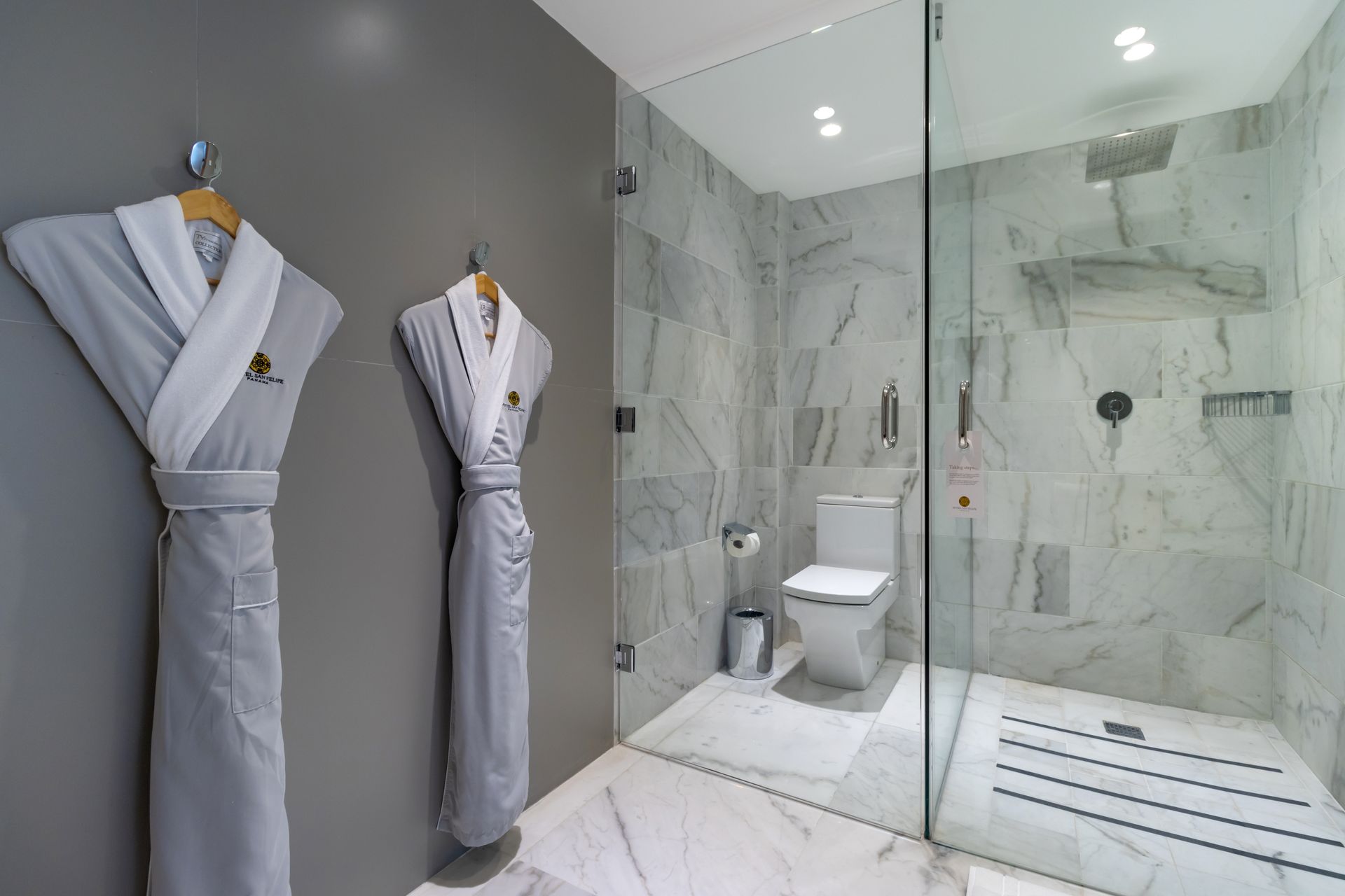 Dos batas grises cuelgan de una pared gris en un baño con azulejos de mármol y ducha de cristal.