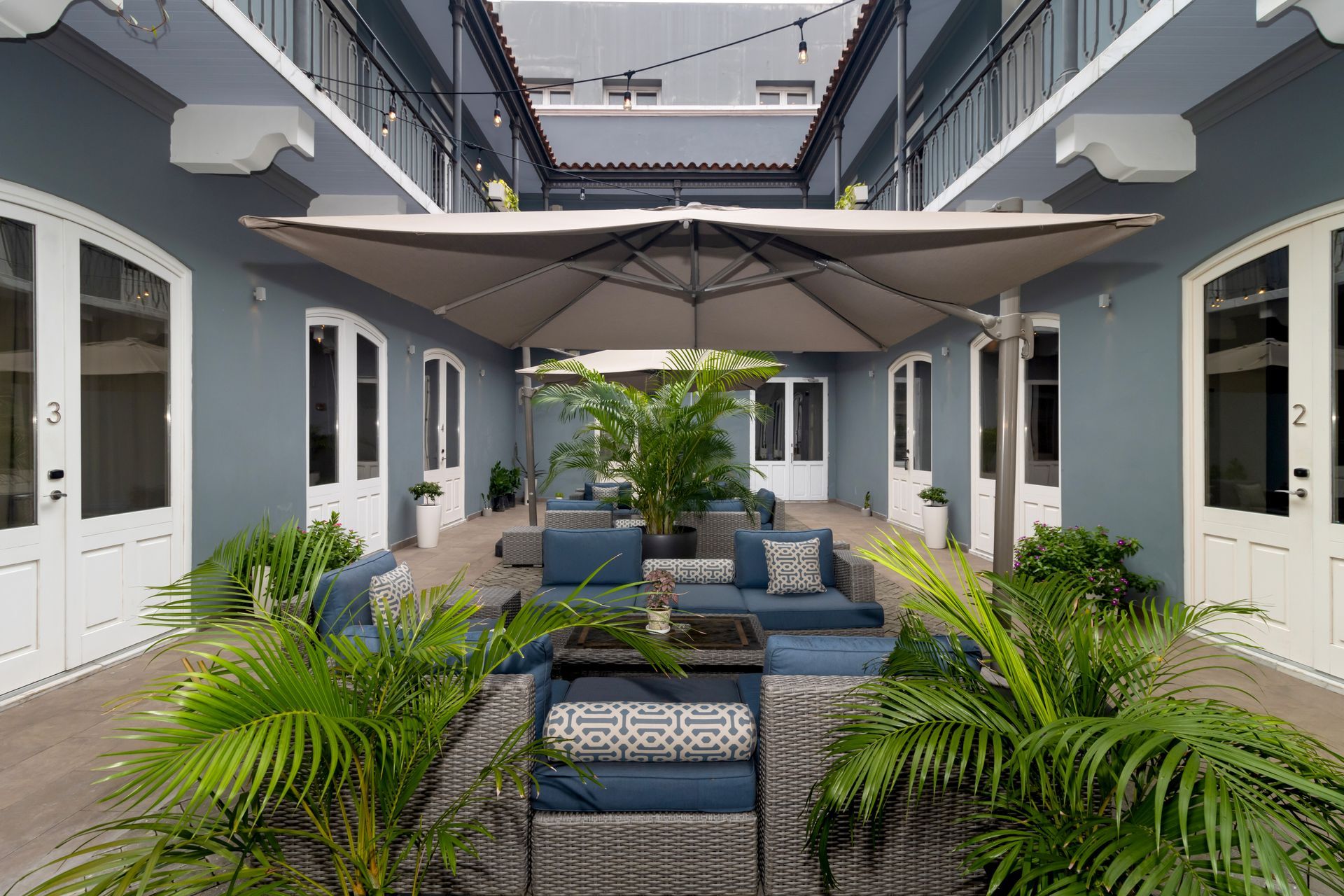 Patio con paredes azules, asientos al aire libre y una gran sombrilla. Palmeras y balcones.