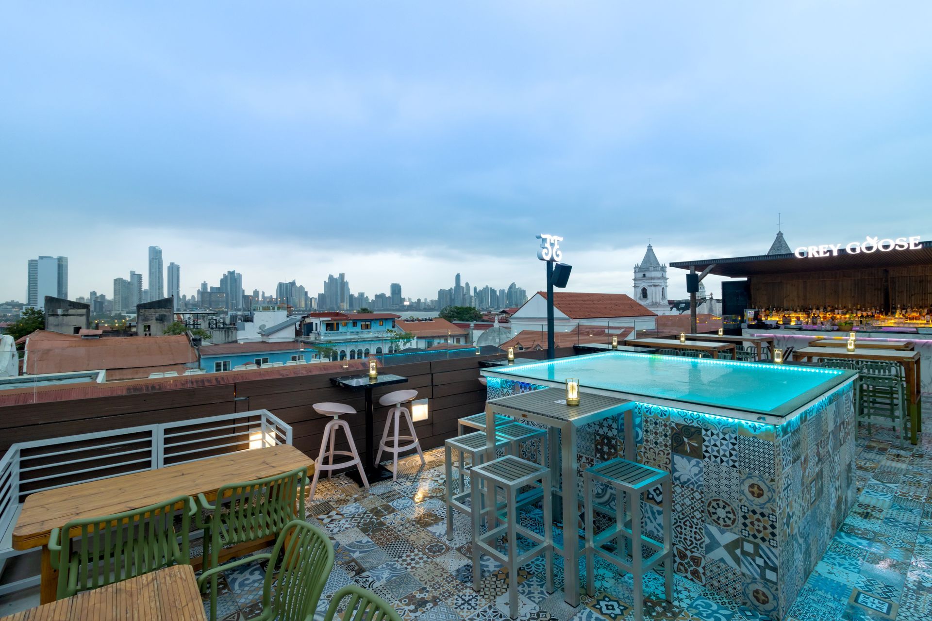 Bar en la azotea con piscina y vistas al horizonte de la ciudad. Se ven mesas, taburetes y una barra.
