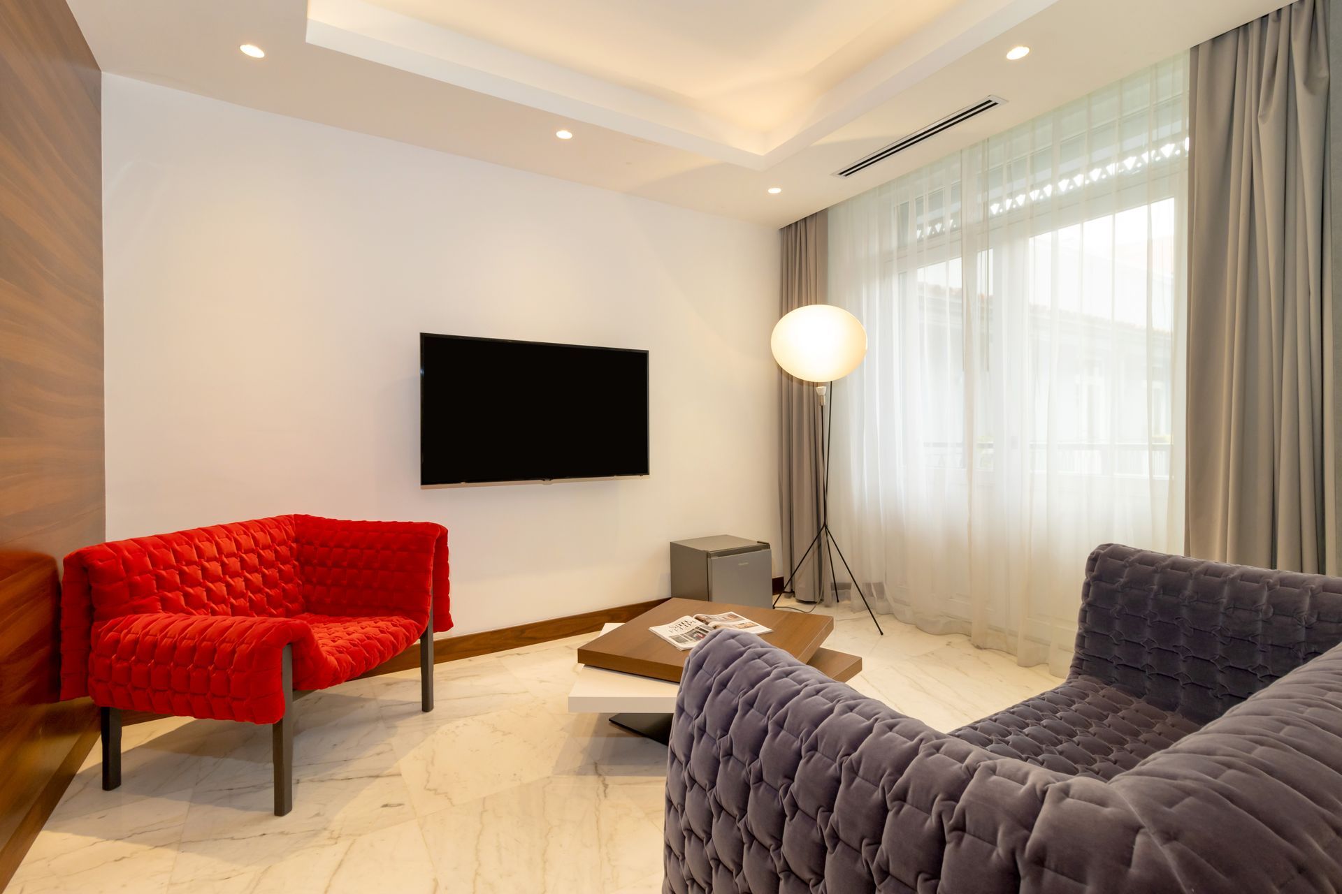 Una sala de estar con un sillón rojo, un sofá gris y un televisor en una pared blanca.