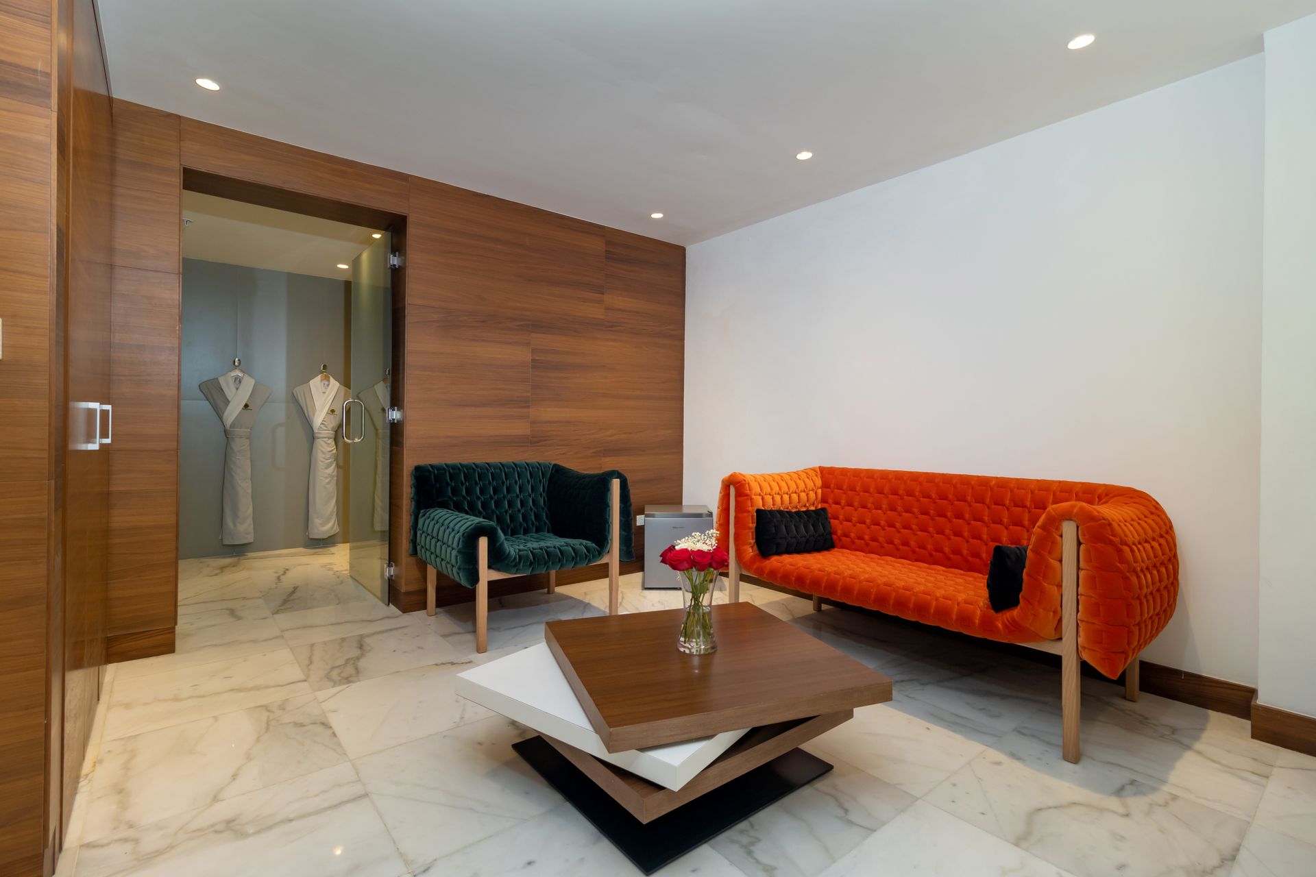 Sala de estar con sofá naranja, sillón verde y detalles en madera, con suelo de mármol. Puerta al baño con albornoces.