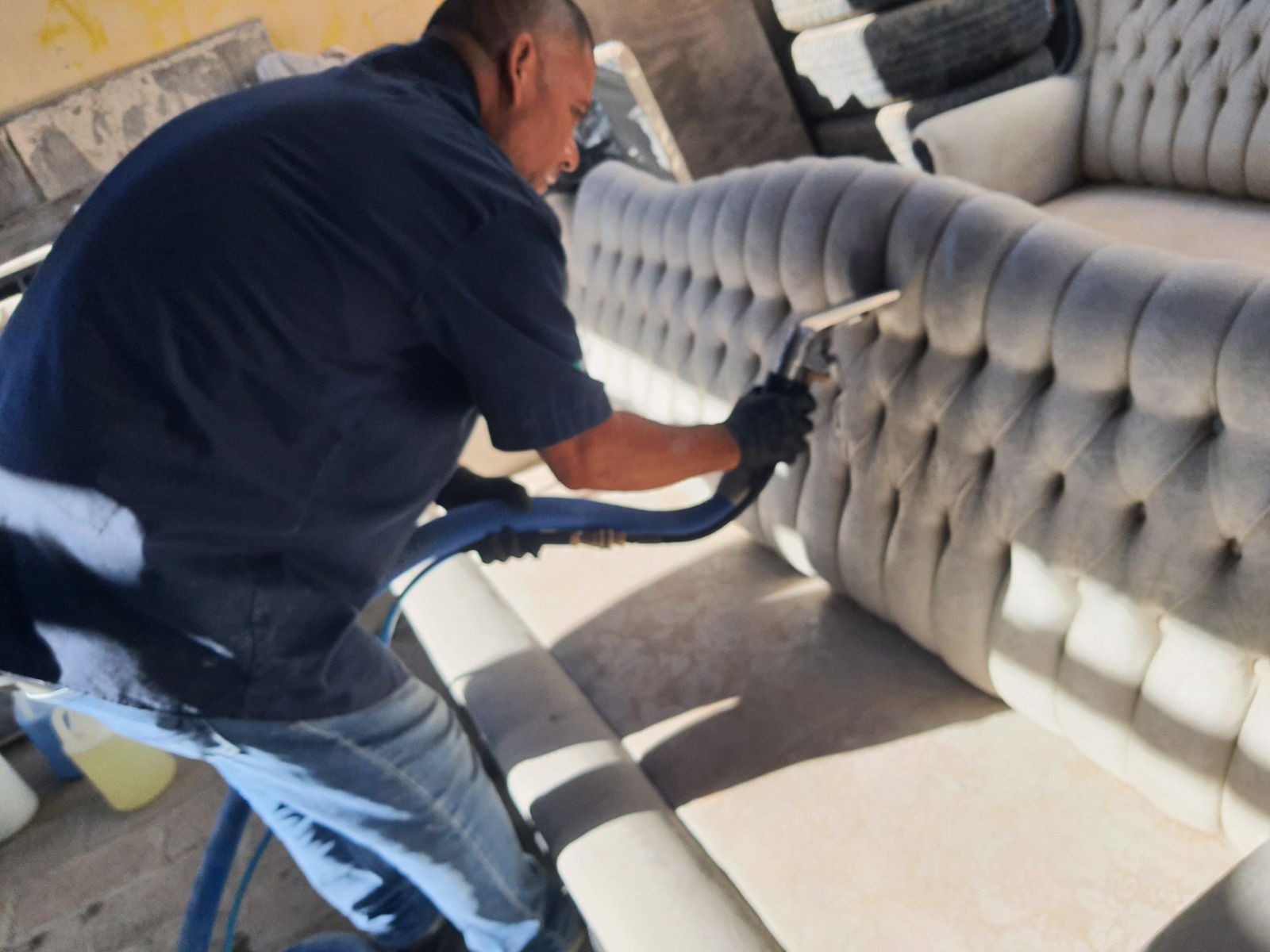 Hombre limpiando un sofá gris con mechones acolchados con una manguera de aspiradora azul en el exterior.