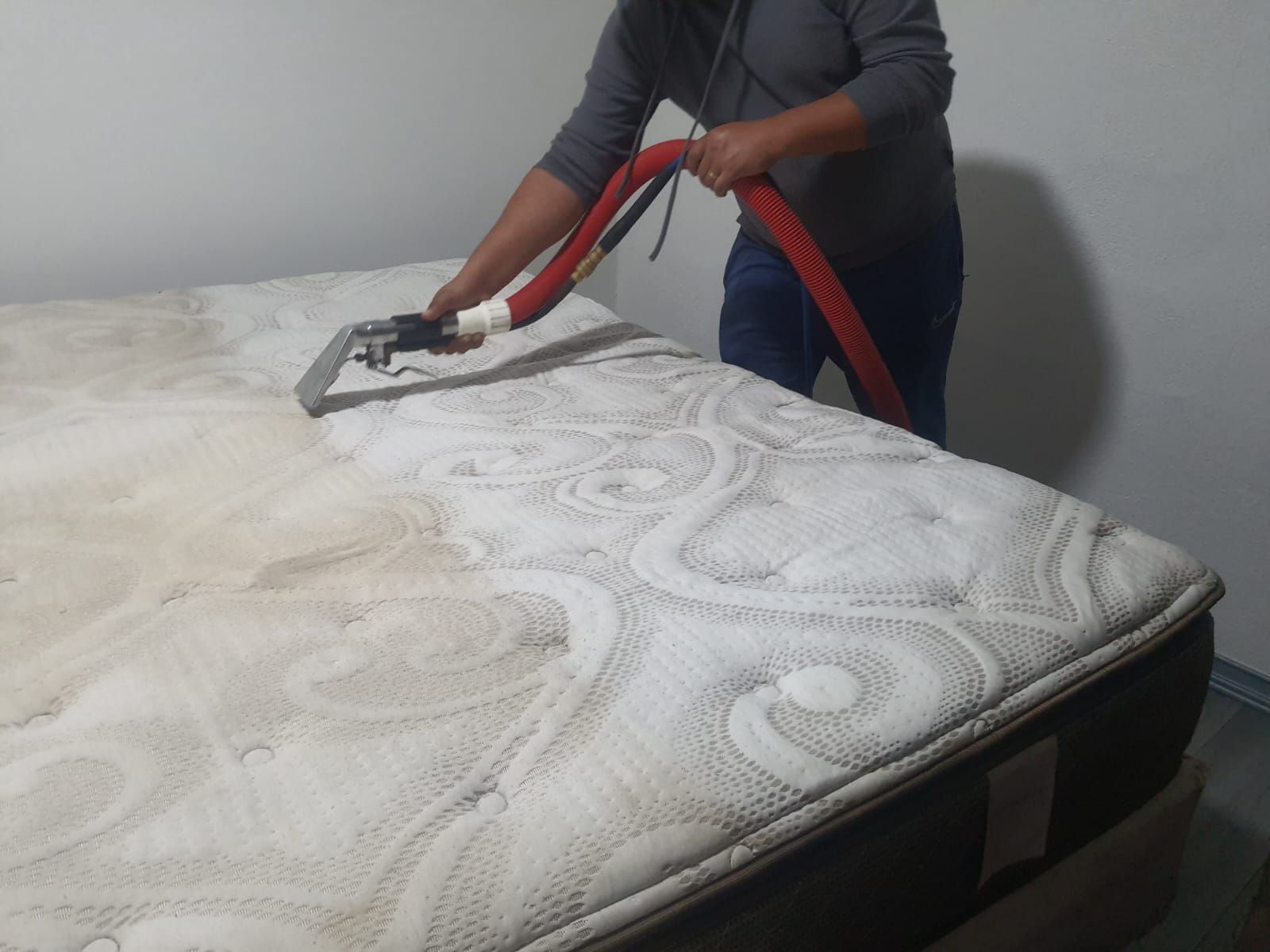 Persona limpiando un colchón con una herramienta de limpieza roja y negra en una habitación de paredes blancas.