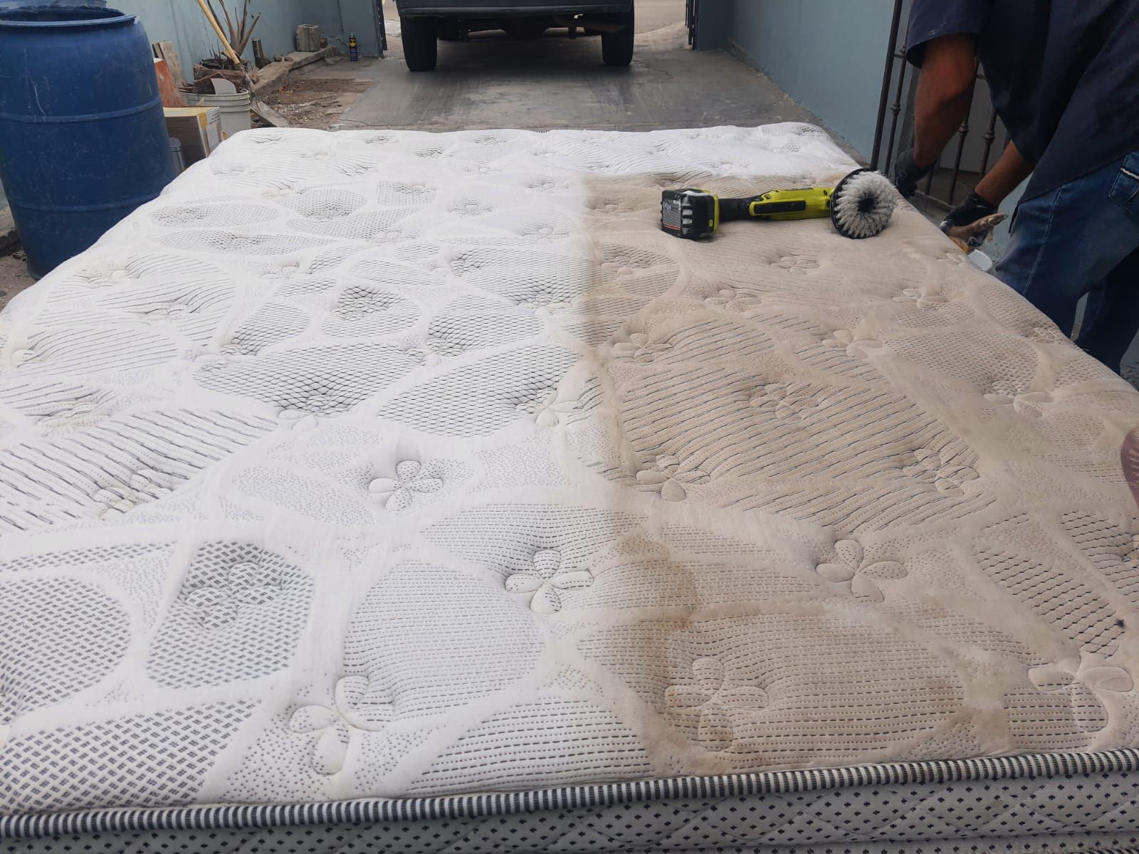 Colchón en proceso de limpieza; un lado parece limpio, el otro sucio. Una persona usa una herramienta de limpieza afuera.