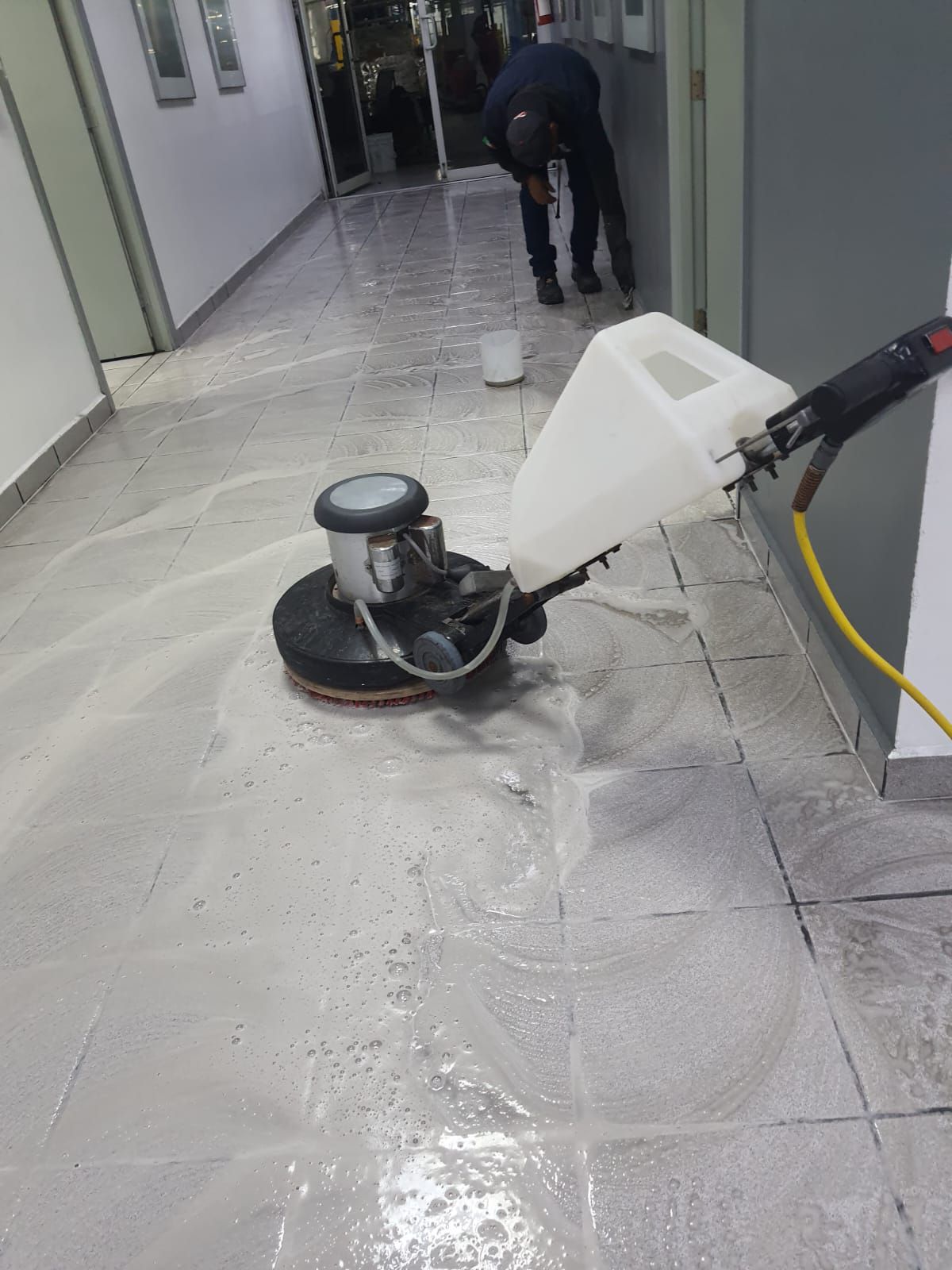 Persona limpiando un piso de baldosas grises con una pulidora en un pasillo.