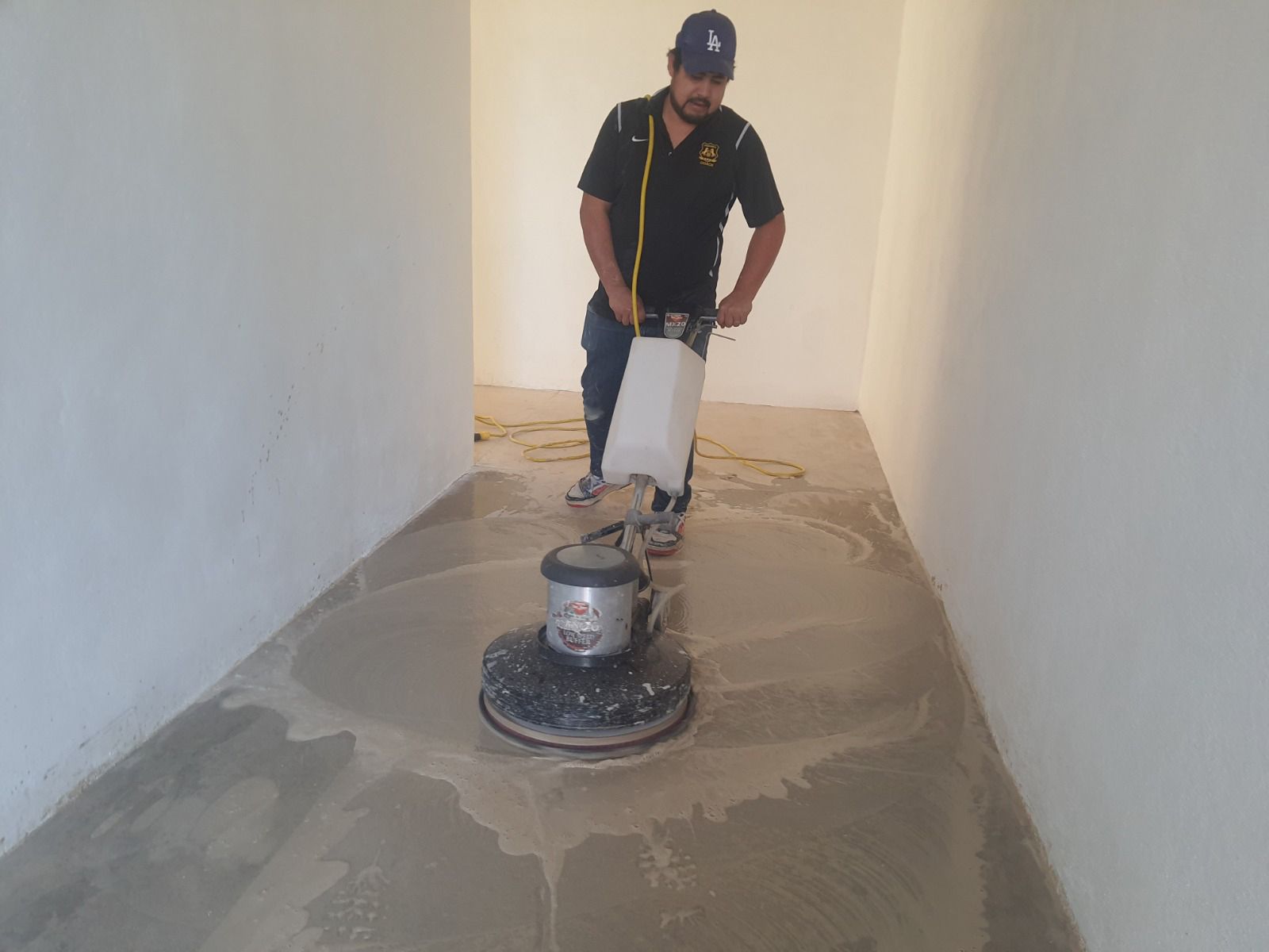 Hombre usando una pulidora de pisos sobre un piso de concreto en una habitación con paredes blancas.