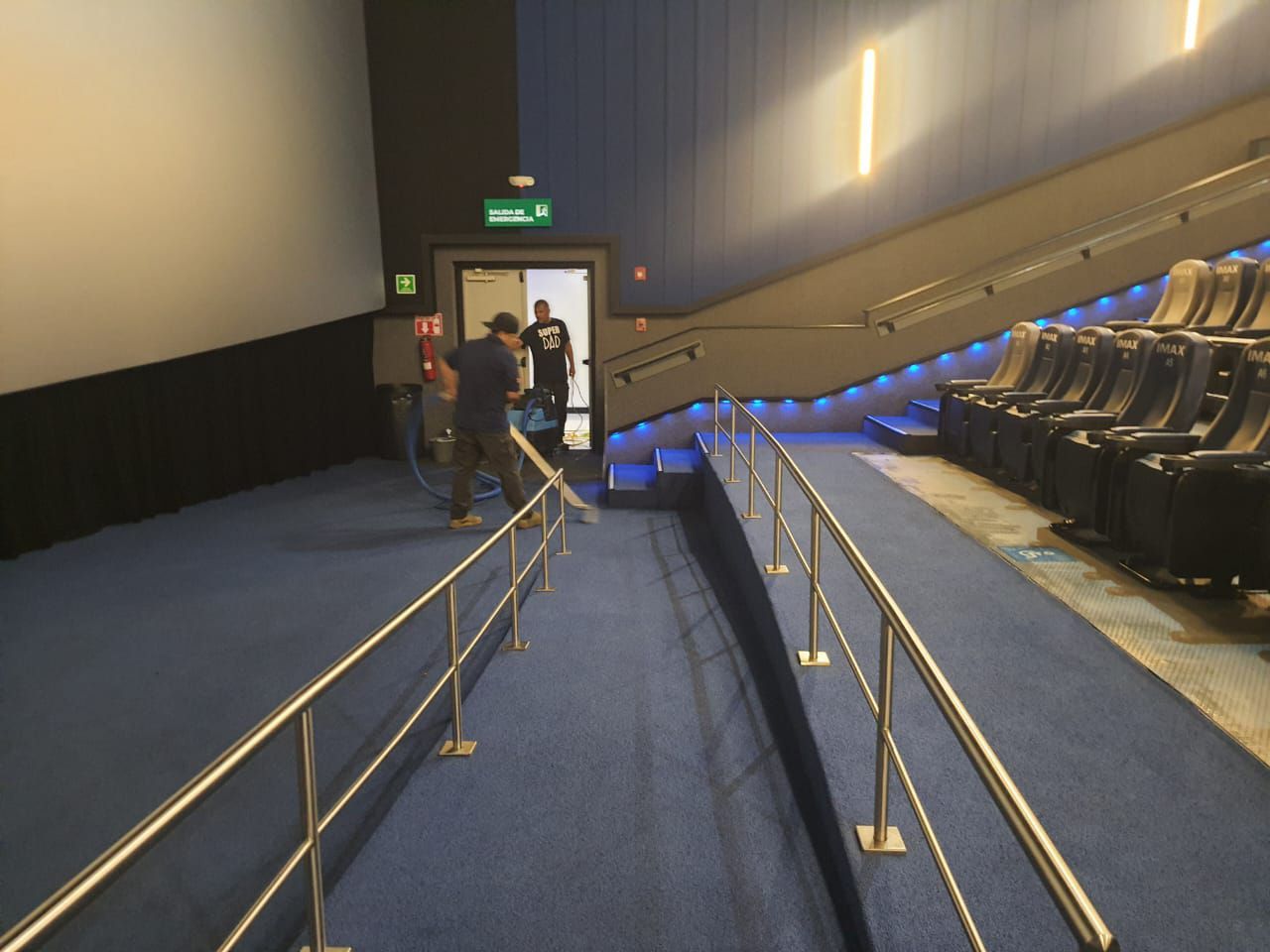 Interior de una sala de cine con rampa para sillas de ruedas, alfombra azul y gente cerca de una salida.