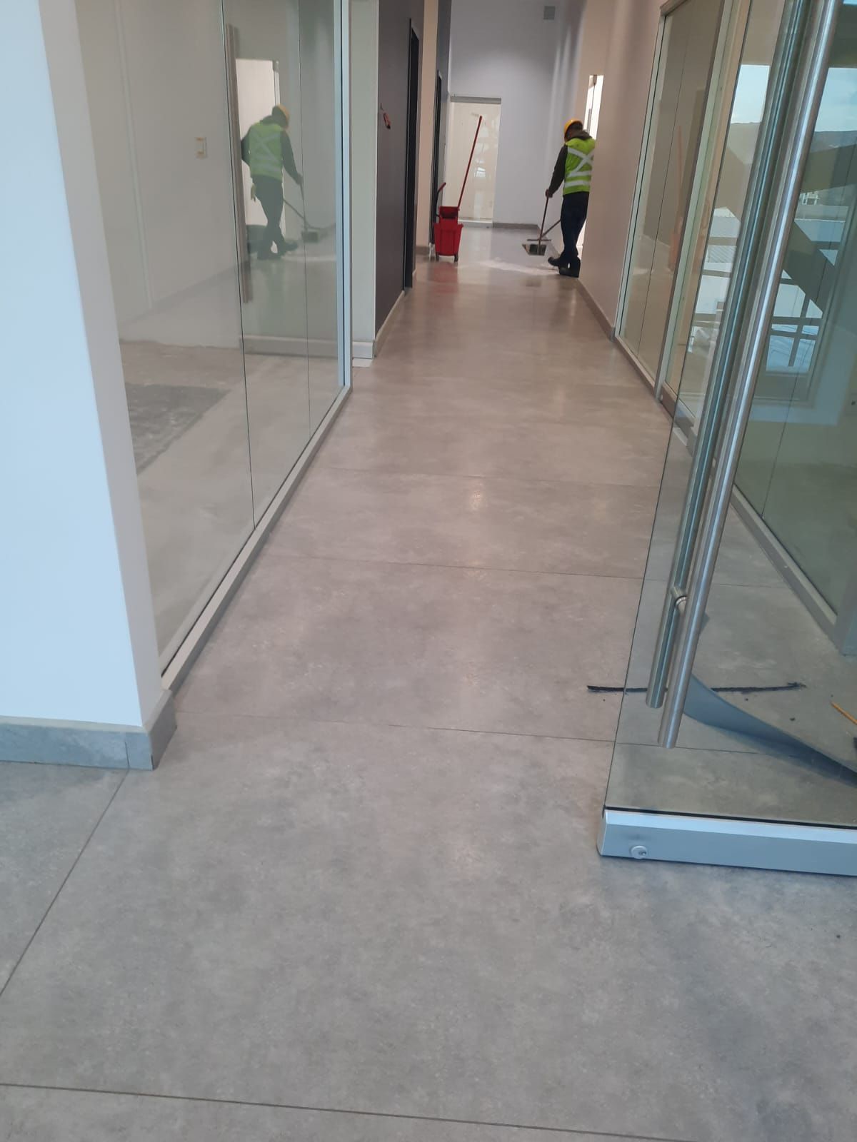 Pasillo largo con suelo de hormigón. Dos personas limpiando. Una barre, la otra se refleja en la pared de cristal.