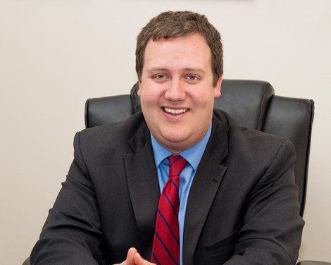 Smiling man in a dark suit and red tie seated at an office desk in a black chair