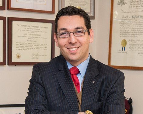 Man in a suit and red tie smiling in an office with framed certificates on the wall